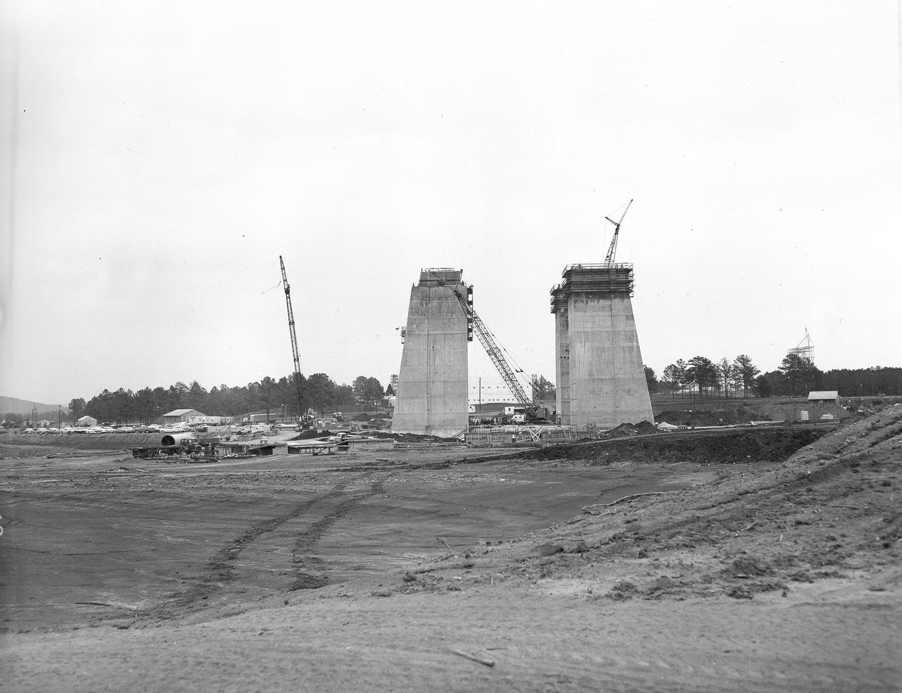 At its founding, the Marshall Space Flight Center (MSFC) inherited the Army’s Jupiter and Redstone test stands, but much larger facilities were needed for the giant stages of the Saturn V. From 1960 to 1964, the existing stands were remodeled and a sizable new test area was developed. The new comprehensive test complex for propulsion and structural dynamics was unique within the nation and the free world, and they remain so today because they were constructed with foresight to meet the future as well as on going needs. Construction of the S-IC Static test stand complex began in 1961 in the west test area of MSFC, and was completed in 1964. The S-IC static test stand was designed to develop and test the 138-ft long and 33-ft diameter Saturn V S-IC first stage, or booster stage, weighing in at 280,000 pounds. Required to hold down the brute force of a 7,500,000-pound thrust produced by 5 F-1 engines, the S-IC static test stand was designed and constructed with the strength of hundreds of tons of steel and 12,000,000 pounds of cement, planted down to bedrock 40 feet below ground level. The foundation walls, constructed with concrete and steel, are 4 feet thick. The base structure consists of four towers with 40-foot-thick walls extending upward 144 feet above ground level. The structure was topped by a crane with a 135-foot boom. With the boom in the upright position, the stand was given an overall height of 405 feet, placing it among the highest structures in Alabama at the time. This photograph taken April 17, 1963, gives a look at the  four tower legs of the S-IC test stand at their completed height.