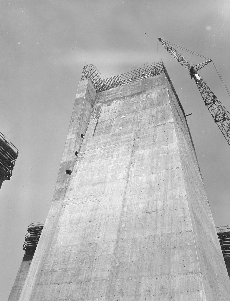 At its founding, the Marshall Space Flight Center (MSFC) inherited the Army’s Jupiter and Redstone test stands, but much larger facilities were needed for the giant stages of the Saturn V. From 1960 to 1964, the existing stands were remodeled and a sizable new test area was developed. The new comprehensive test complex for propulsion and structural dynamics was unique within the nation and the free world, and they remain so today because they were constructed with foresight to meet the future as well as on going needs. Construction of the S-IC Static test stand complex began in 1961 in the west test area of MSFC, and was completed in 1964. The S-IC static test stand was designed to develop and test the 138-ft long and 33-ft diameter Saturn V S-IC first stage, or booster stage, weighing in at 280,000 pounds. Required to hold down the brute force of a 7,500,000-pound thrust produced by 5 F-1 engines, the S-IC static test stand was designed and constructed with the strength of hundreds of tons of steel and 12,000,000 pounds of cement, planted down to bedrock 40 feet below ground level. The foundation walls, constructed with concrete and steel, are 4 feet thick. The base structure consists of four towers with 40-foot-thick walls extending upward 144 feet above ground level. The structure was topped by a crane with a 135-foot boom. With the boom in the upright position, the stand was given an overall height of 405 feet, placing it among the highest structures in Alabama at the time. This photograph, taken from ground level on May 7, 1963, gives a close look at one of the four towers legs of the S-IC test stand nearing its completed height.