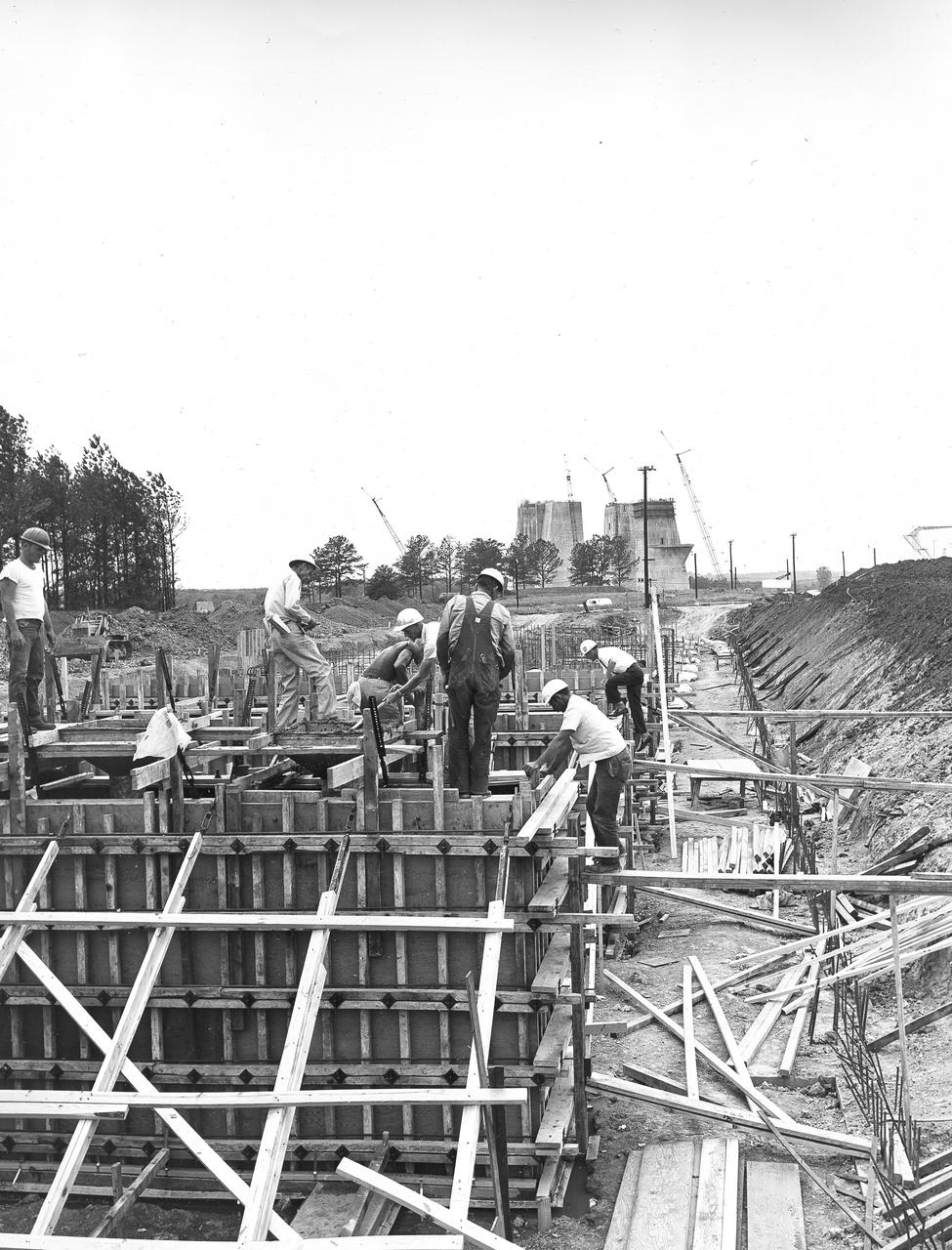 At its founding, the Marshall Space Flight Center (MSFC) inherited the Army’s Jupiter and Redstone test stands, but much larger facilities were needed for the giant stages of the Saturn V. From 1960 to 1964, the existing stands were remodeled and a sizable new test area was developed. The new comprehensive test complex for propulsion and structural dynamics was unique within the nation and the free world, and they remain so today because they were constructed with foresight to meet the future as well as on going needs. Construction of the S-IC Static test stand complex began in 1961 in the west test area of MSFC, and was completed in 1964. The S-IC static test stand was designed to develop and test the 138-ft long and 33-ft diameter Saturn V S-IC first stage, or booster stage, weighing in at 280,000 pounds. Required to hold down the brute force of a 7,500,000-pound thrust produced by 5 F-1 engines, the S-IC static test stand was designed and constructed with the strength of hundreds of tons of steel and 12,000,000 pounds of cement, planted down to bedrock 40 feet below ground level. The foundation walls, constructed with concrete and steel, are 4 feet thick. The base structure consists of four towers with 40-foot-thick walls extending upward 144 feet above ground level. The structure was topped by a crane with a 135-foot boom. With the boom in the upright position, the stand was given an overall height of 405 feet, placing it among the highest structures in Alabama at the time. In addition to the stand itself, related facilities were constructed during this time. Built directly east of the test stand was the Block House, which served as the control center for the test stand. The two were connected by a narrow access tunnel which housed the cables for the controls. Again to the east, just south of the Block House, was a newly constructed Pump House. Its function was to provide water to the stand to prevent melting damage during testing. The water was sprayed through small holes in the stand’s 1900 ton water deflector at the rate of 320,000 gallons per minute. In this photo, taken May 22, 1963, the Pump House is undergoing construction.