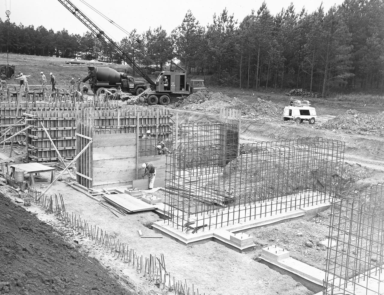 At its founding, the Marshall Space Flight Center (MSFC) inherited the Army’s Jupiter and Redstone test stands, but much larger facilities were needed for the giant stages of the Saturn V. From 1960 to 1964, the existing stands were remodeled and a sizable new test area was developed. The new comprehensive test complex for propulsion and structural dynamics was unique within the nation and the free world, and they remain so today because they were constructed with foresight to meet the future as well as on going needs. Construction of the S-IC Static test stand complex began in 1961 in the west test area of MSFC, and was completed in 1964. The S-IC static test stand was designed to develop and test the 138-ft long and 33-ft diameter Saturn V S-IC first stage, or booster stage, weighing in at 280,000 pounds. Required to hold down the brute force of a 7,500,000-pound thrust produced by 5 F-1 engines, the S-IC static test stand was designed and constructed with the strength of hundreds of tons of steel and 12,000,000 pounds of cement, planted down to bedrock 40 feet below ground level. The foundation walls, constructed with concrete and steel, are 4 feet thick. The base structure consists of four towers with 40-foot-thick walls extending upward 144 feet above ground level. The structure was topped by a crane with a 135-foot boom. With the boom in the upright position, the stand was given an overall height of 405 feet, placing it among the highest structures in Alabama at the time. In addition to the stand itself, related facilities were constructed during this time. Built directly east of the test stand was the Block House, which served as the control center for the test stand. The two were connected by a narrow access tunnel which housed the cables for the controls. Again to the east, just south of the Block House, was a newly constructed Pump House. Its function was to provide water to the stand to prevent melting damage during testing. The water was sprayed through small holes in the stand’s 1900 ton water deflector at the rate of 320,000 gallons per minute. In this photo, taken May 22, 1963, the Pump House is undergoing construction.
