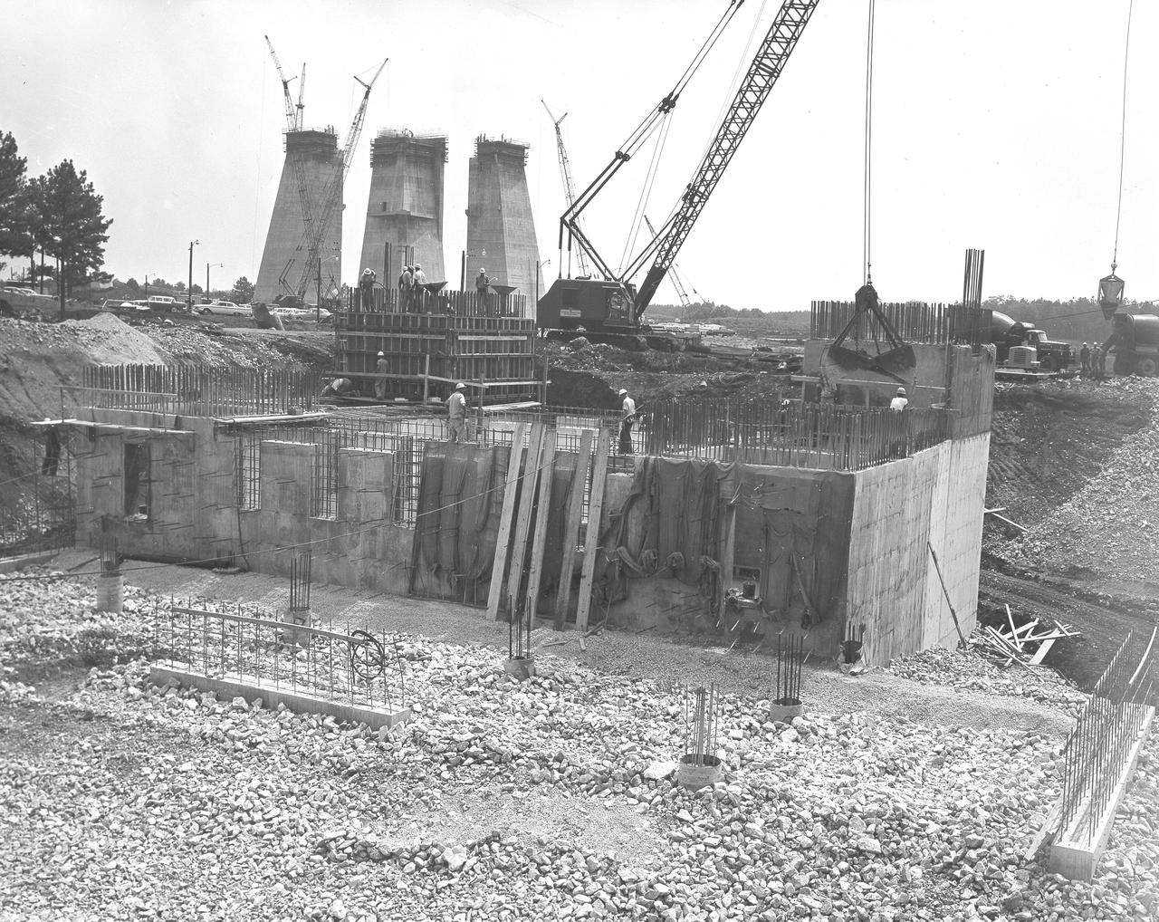 At its founding, the Marshall Space Flight Center (MSFC) inherited the Army’s Jupiter and Redstone test stands, but much larger facilities were needed for the giant stages of the Saturn V. From 1960 to 1964, the existing stands were remodeled and a sizable new test area was developed. The new comprehensive test complex for propulsion and structural dynamics was unique within the nation and the free world, and they remain so today because they were constructed with foresight to meet the future as well as on going needs. Construction of the S-IC Static test stand complex began in 1961 in the west test area of MSFC, and was completed in 1964. The S-IC static test stand was designed to develop and test the 138-ft long and 33-ft diameter Saturn V S-IC first stage, or booster stage, weighing in at 280,000 pounds. Required to hold down the brute force of a 7,500,000-pound thrust produced by 5 F-1 engines, the S-IC static test stand was designed and constructed with the strength of hundreds of tons of steel and 12,000,000 pounds of cement, planted down to bedrock 40 feet below ground level. The foundation walls, constructed with concrete and steel, are 4 feet thick. The base structure consists of four towers with 40-foot-thick walls extending upward 144 feet above ground level. The structure was topped by a crane with a 135-foot boom. With the boom in the upright position, the stand was given an overall height of 405 feet, placing it among the highest structures in Alabama at the time. In addition to the stand itself, related facilities were constructed during this time. North of the massive S-IC test stand, the F-1 Engine test stand was built. Designed to assist in the development of the F-1 Engine, the F-1 test stand is a vertical engine firing test stand, 239 feet in elevation and 4,600 square feet in area at the base. Capability was provided for static firing of 1.5 million pounds of thrust using liquid oxygen and kerosene. Like the S-IC stand, the foundation of the F-1 stand is keyed into the bedrock approximately 40 feet below grade. This photo depicts the construction of the F-1 test stand as of June 24, 1963. Two if its four tower legs are underway.