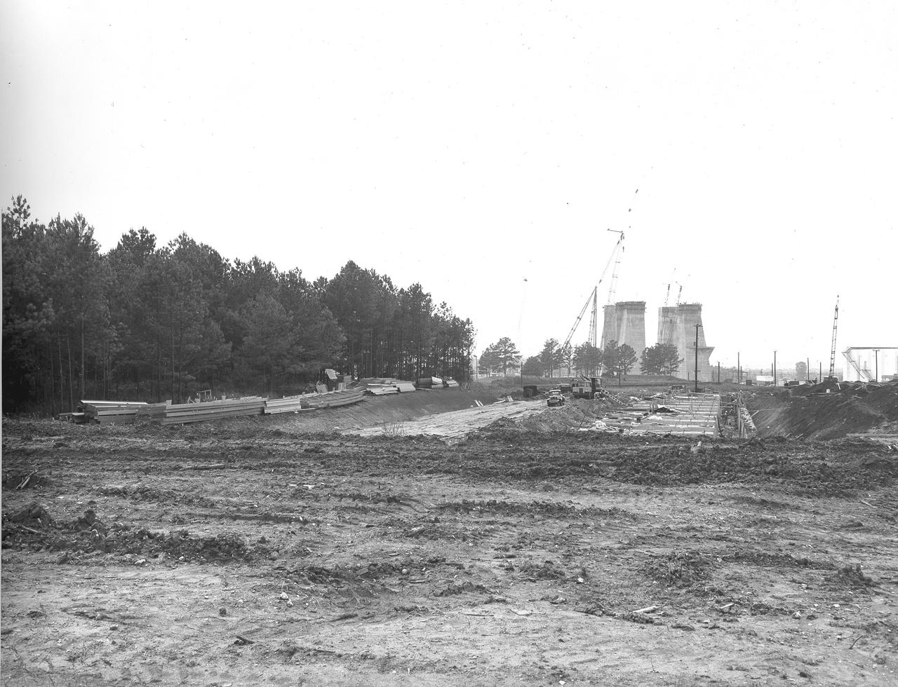 At its founding, the Marshall Space Flight Center (MSFC) inherited the Army’s Jupiter and Redstone test stands, but much larger facilities were needed for the giant stages of the Saturn V. From 1960 to 1964, the existing stands were remodeled and a sizable new test area was developed. The new comprehensive test complex for propulsion and structural dynamics was unique within the nation and the free world, and they remain so today because they were constructed with foresight to meet the future as well as on going needs. Construction of the S-IC Static test stand complex began in 1961 in the west test area of MSFC, and was completed in 1964. The S-IC static test stand was designed to develop and test the 138-ft long and 33-ft diameter Saturn V S-IC first stage, or booster stage, weighing in at 280,000 pounds. Required to hold down the brute force of a 7,500,000-pound thrust produced by 5 F-1 engines, the S-IC static test stand was designed and constructed with the strength of hundreds of tons of steel and 12,000,000 pounds of cement, planted down to bedrock 40 feet below ground level. The foundation walls, constructed with concrete and steel, are 4 feet thick. The base structure consists of four towers with 40-foot-thick walls extending upward 144 feet above ground level. The structure was topped by a crane with a 135-foot boom. With the boom in the upright position, the stand was given an overall height of 405 feet, placing it among the highest structures in Alabama at the time. In addition to the stand itself, related facilities were constructed during this time. Built directly east of the test stand was the Block House, which served as the control center for the test stand. The two were connected by a narrow access tunnel which housed the cables for the controls. Again to the east, just south of the Block House, was a newly constructed Pump House. Its function was to provide water to the stand to prevent melting damage during testing. The water was sprayed through small holes in the stand’s 1900 ton water deflector at the rate of 320,000 gallons per minute. In this photo, taken July 3, 1963, the Pump House is undergoing construction and has reached ground level.