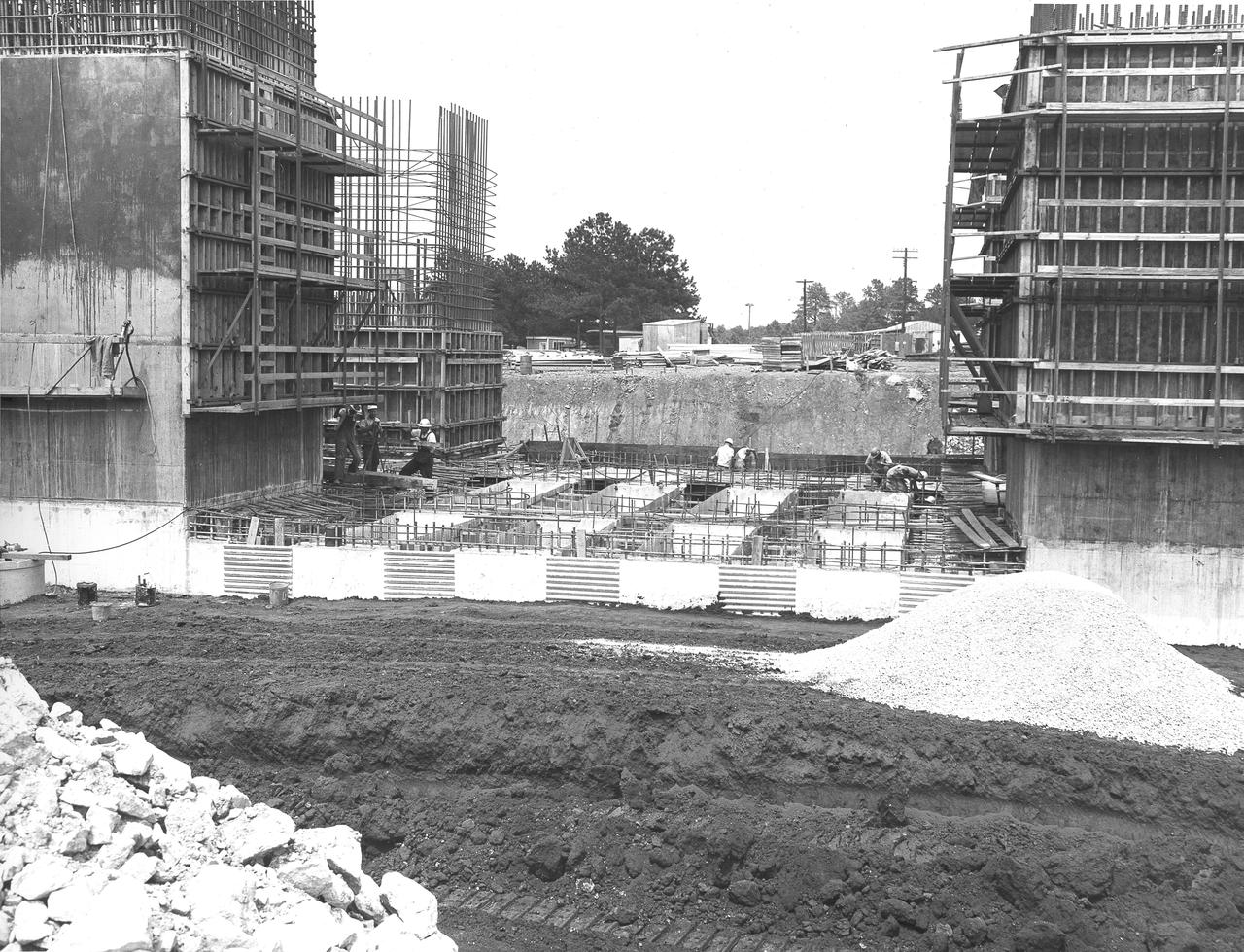 At its founding, the Marshall Space Flight Center (MSFC) inherited the Army’s Jupiter and Redstone test stands, but much larger facilities were needed for the giant stages of the Saturn V. From 1960 to 1964, the existing stands were remodeled and a sizable new test area was developed. The new comprehensive test complex for propulsion and structural dynamics was unique within the nation and the free world, and they remain so today because they were constructed with foresight to meet the future as well as on going needs. Construction of the S-IC Static test stand complex began in 1961 in the west test area of MSFC, and was completed in 1964. The S-IC static test stand was designed to develop and test the 138-ft long and 33-ft diameter Saturn V S-IC first stage, or booster stage, weighing in at 280,000 pounds. Required to hold down the brute force of a 7,500,000-pound thrust produced by 5 F-1 engines, the S-IC static test stand was designed and constructed with the strength of hundreds of tons of steel and 12,000,000 pounds of cement, planted down to bedrock 40 feet below ground level. The foundation walls, constructed with concrete and steel, are 4 feet thick. The base structure consists of four towers with 40-foot-thick walls extending upward 144 feet above ground level. The structure was topped by a crane with a 135-foot boom. With the boom in the upright position, the stand was given an overall height of 405 feet, placing it among the highest structures in Alabama at the time. In addition to the stand itself, related facilities were constructed during this time. North of the massive S-IC test stand, the F-1 Engine test stand was built. Designed to assist in the development of the F-1 Engine, the F-1 test stand is a vertical engine firing test stand, 239 feet in elevation and 4,600 square feet in area at the base. Capability was provided for static firing of 1.5 million pounds of thrust using liquid oxygen and kerosene. Like the S-IC stand, the foundation of the F-1 stand is keyed into the bedrock approximately 40 feet below grade. This photo depicts the construction of the F-1 test stand as of July 3, 1963. All four of its tower legs are well underway.