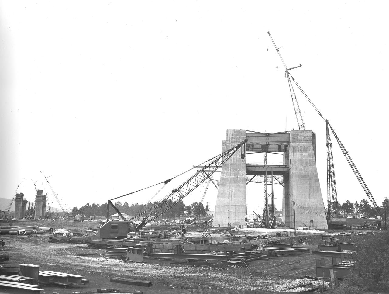 At its founding, the Marshall Space Flight Center (MSFC) inherited the Army’s Jupiter and Redstone test stands, but much larger facilities were needed for the giant stages of the Saturn V. From 1960 to 1964, the existing stands were remodeled and a sizable new test area was developed. The new comprehensive test complex for propulsion and structural dynamics was unique within the nation and the free world, and they remain so today because they were constructed with foresight to meet the future as well as on going needs. Construction of the S-IC Static test stand complex began in 1961 in the west test area of MSFC, and was completed in 1964. The S-IC static test stand was designed to develop and test the 138-ft long and 33-ft diameter Saturn V S-IC first stage, or booster stage, weighing in at 280,000 pounds. Required to hold down the brute force of a 7,500,000-pound thrust produced by 5 F-1 engines, the S-IC static test stand was designed and constructed with the strength of hundreds of tons of steel and 12,000,000 pounds of cement, planted down to bedrock 40 feet below ground level. The foundation walls, constructed with concrete and steel, are 4 feet thick. The base structure consists of four towers with 40-foot-thick walls extending upward 144 feet above ground level. The structure was topped by a crane with a 135-foot boom. With the boom in the upright position, the stand was given an overall height of 405 feet, placing it among the highest structures in Alabama at the time. In addition to the stand itself, related facilities were constructed during this time. Built to the east was a newly constructed Pump House. Its function was to provide water to the stand to prevent melting damage during testing. The water was sprayed through small holes in the stand’s 1900 ton flame deflector at the rate of 320,000 gallons per minute. In this photo, taken August 12, 1963, the S-IC stand has received some of its internal components.  Directly in the center is the framework that houses the flame deflector. The F-1 test stand, designed and built to test a single F-1 engine, can be seen on the left side of the photo.