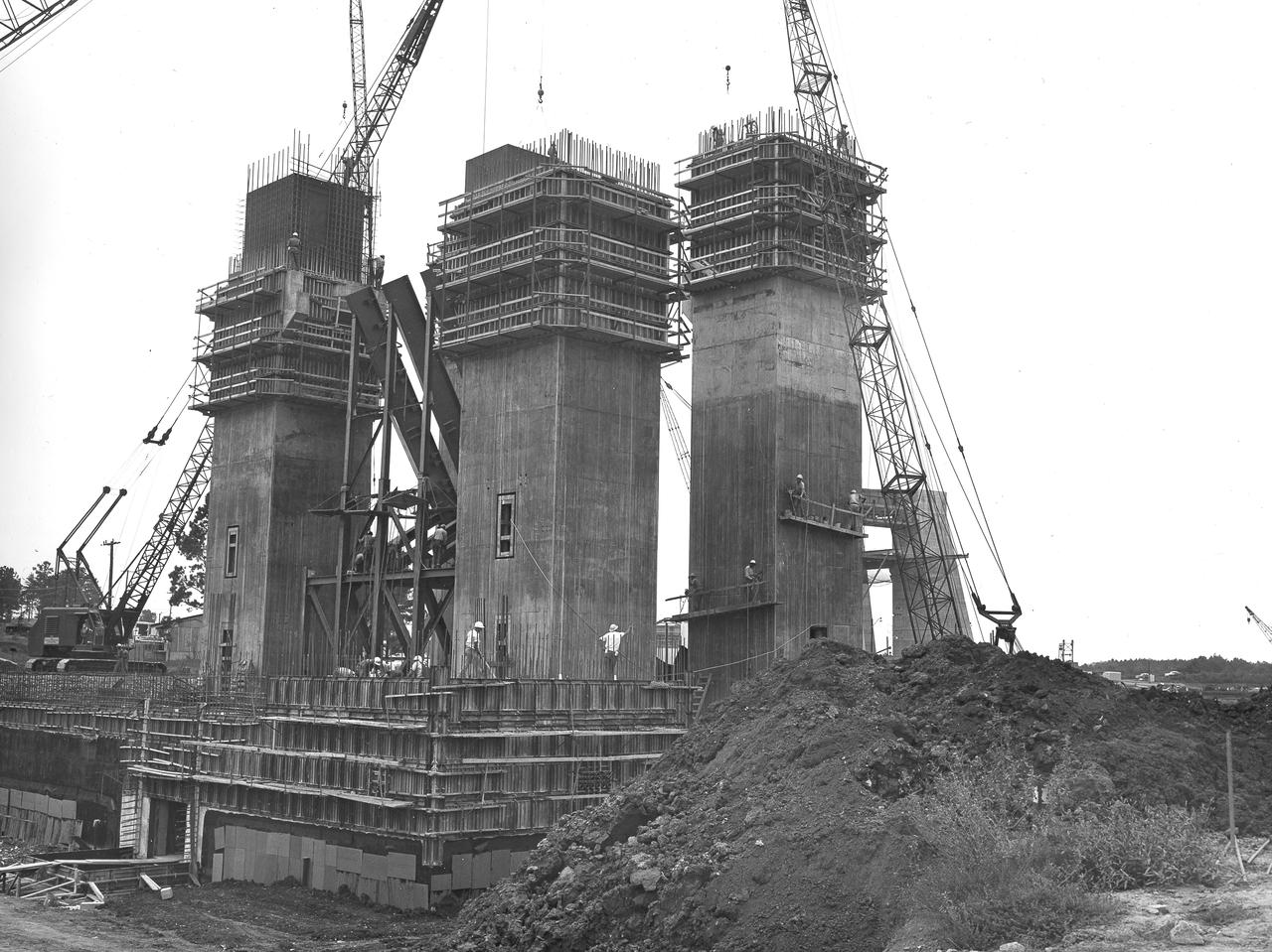 At its founding, the Marshall Space Flight Center (MSFC) inherited the Army’s Jupiter and Redstone test stands, but much larger facilities were needed for the giant stages of the Saturn V. From 1960 to 1964, the existing stands were remodeled and a sizable new test area was developed. The new comprehensive test complex for propulsion and structural dynamics was unique within the nation and the free world, and they remain so today because they were constructed with foresight to meet the future as well as on going needs. Construction of the S-IC Static test stand complex began in 1961 in the west test area of MSFC, and was completed in 1964. The S-IC static test stand was designed to develop and test the 138-ft long and 33-ft diameter Saturn V S-IC first stage, or booster stage, weighing in at 280,000 pounds. Required to hold down the brute force of a 7,500,000-pound thrust produced by 5 F-1 engines, the S-IC static test stand was designed and constructed with the strength of hundreds of tons of steel and 12,000,000 pounds of cement, planted down to bedrock 40 feet below ground level. The foundation walls, constructed with concrete and steel, are 4 feet thick. The base structure consists of four towers with 40-foot-thick walls extending upward 144 feet above ground level. The structure was topped by a crane with a 135-foot boom. With the boom in the upright position, the stand was given an overall height of 405 feet, placing it among the highest structures in Alabama at the time. In addition to the stand itself, related facilities were constructed during this time. North of the massive S-IC test stand, the F-1 Engine test stand was built. Designed to assist in the development of the F-1 Engine, the F-1 test stand is a vertical engine firing test stand, 239 feet in elevation and 4,600 square feet in area at the base. Capability was provided for static firing of 1.5 million pounds of thrust using liquid oxygen and kerosene. Like the S-IC stand, the foundation of the F-1 stand is keyed into the bedrock approximately 40 feet below grade. This photo depicts the construction of the F-1 test stand as of August 13, 1963. All four of its tower legs are well underway into the skyline.