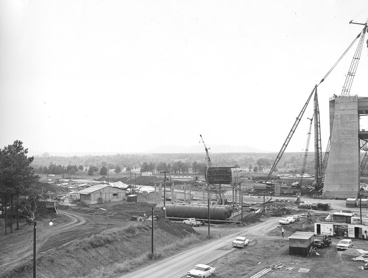 At its founding, the Marshall Space Flight Center (MSFC) inherited the Army’s Jupiter and Redstone test stands, but much larger facilities were needed for the giant stages of the Saturn V. From 1960 to 1964, the existing stands were remodeled and a sizable new test area was developed. The new comprehensive test complex for propulsion and structural dynamics was unique within the nation and the free world, and they remain so today because they were constructed with foresight to meet the future as well as on going needs. Construction of the S-IC Static test stand complex began in 1961 in the west test area of MSFC, and was completed in 1964. The S-IC static test stand was designed to develop and test the 138-ft long and 33-ft diameter Saturn V S-IC first stage, or booster stage, weighing in at 280,000 pounds. Required to hold down the brute force of a 7,500,000-pound thrust produced by 5 F-1 engines, the S-IC static test stand was designed and constructed with the strength of hundreds of tons of steel and 12,000,000 pounds of cement, planted down to bedrock 40 feet below ground level. The foundation walls, constructed with concrete and steel, are 4 feet thick. The base structure consists of four towers with 40-foot-thick walls extending upward 144 feet above ground level. The structure was topped by a crane with a 135-foot boom. With the boom in the upright position, the stand was given an overall height of 405 feet, placing it among the highest structures in Alabama at the time. In addition to the stand itself, related facilities were constructed during this time. In the center portion of this photograph, taken September 5, 1963, the spherical hydrogen storage tanks are being constructed.  One of the massive tower legs of the S-IC test stand is visible to the far right.