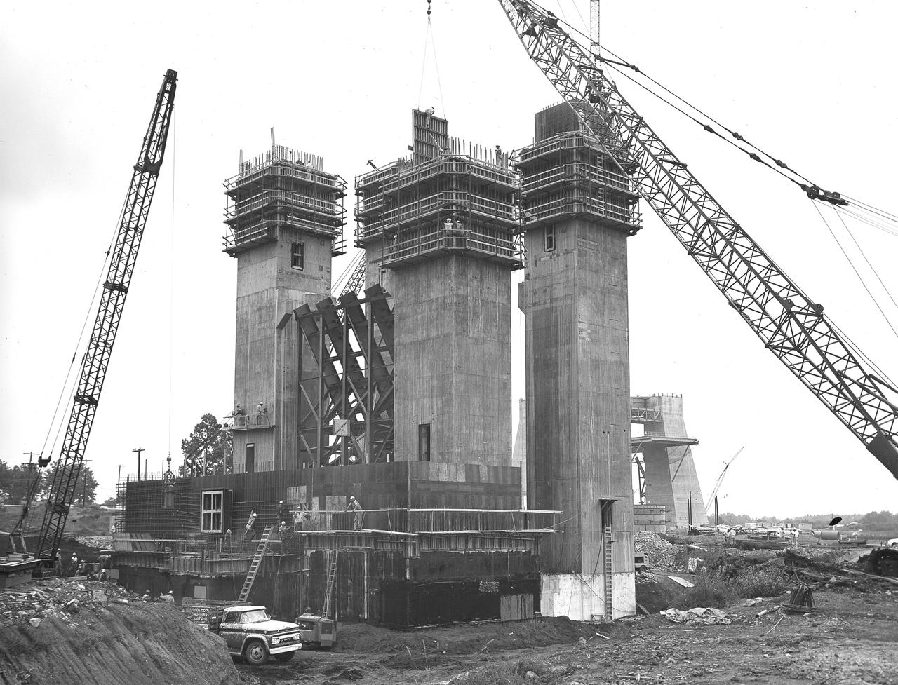 At its founding, the Marshall Space Flight Center (MSFC) inherited the Army’s Jupiter and Redstone test stands, but much larger facilities were needed for the giant stages of the Saturn V. From 1960 to 1964, the existing stands were remodeled and a sizable new test area was developed. The new comprehensive test complex for propulsion and structural dynamics was unique within the nation and the free world, and they remain so today because they were constructed with foresight to meet the future as well as on going needs. Construction of the S-IC Static test stand complex began in 1961 in the west test area of MSFC, and was completed in 1964. The S-IC static test stand was designed to develop and test the 138-ft long and 33-ft diameter Saturn V S-IC first stage, or booster stage, weighing in at 280,000 pounds. Required to hold down the brute force of a 7,500,000-pound thrust produced by 5 F-1 engines, the S-IC static test stand was designed and constructed with the strength of hundreds of tons of steel and 12,000,000 pounds of cement, planted down to bedrock 40 feet below ground level. The foundation walls, constructed with concrete and steel, are 4 feet thick. The base structure consists of four towers with 40-foot-thick walls extending upward 144 feet above ground level. The structure was topped by a crane with a 135-foot boom. With the boom in the upright position, the stand was given an overall height of 405 feet, placing it among the highest structures in Alabama at the time. In addition to the stand itself, related facilities were constructed during this time. North of the massive S-IC test stand, the F-1 Engine test stand was built. Designed to assist in the development of the F-1 Engine, the F-1 test stand is a vertical engine firing test stand, 239 feet in elevation and 4,600 square feet in area at the base. Capability was provided for static firing of 1.5 million pounds of thrust using liquid oxygen and kerosene. Like the S-IC stand, the foundation of the F-1 stand is keyed into the bedrock approximately 40 feet below grade. This photo depicts the construction of the F-1 test stand as of September 5, 1963. 