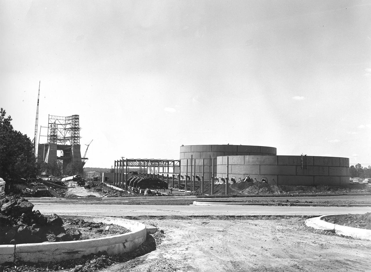 At its founding, the Marshall Space Flight Center (MSFC) inherited the Army’s Jupiter and Redstone test stands, but much larger facilities were needed for the giant stages of the Saturn V. From 1960 to 1964, the existing stands were remodeled and a sizable new test area was developed. The new comprehensive test complex for propulsion and structural dynamics was unique within the nation and the free world, and they remain so today because they were constructed with foresight to meet the future as well as on going needs. Construction of the S-IC Static test stand complex began in 1961 in the west test area of MSFC, and was completed in 1964. The S-IC static test stand was designed to develop and test the 138-ft long and 33-ft diameter Saturn V S-IC first stage, or booster stage, weighing in at 280,000 pounds. Required to hold down the brute force of a 7,500,000-pound thrust produced by 5 F-1 engines, the S-IC static test stand was designed and constructed with the strength of hundreds of tons of steel and 12,000,000 pounds of cement, planted down to bedrock 40 feet below ground level. The foundation walls, constructed with concrete and steel, are 4 feet thick. The base structure consists of four towers with 40-foot-thick walls extending upward 144 feet above ground level. The structure was topped by a crane with a 135-foot boom. With the boom in the upright position, the stand was given an overall height of 405 feet, placing it among the highest structures in Alabama at the time. In addition to the stand itself, related facilities were constructed during this time. Built to the northeast of the stand was a newly constructed Pump House. Its function was to provide water to the stand to prevent melting damage during testing. The water was sprayed through small holes in the stand’s 1900 ton flame deflector at the rate of 320,000 gallons per minute. This photograph, taken September 25, 1963, depicts the construction progress of the Pump House and massive round water tanks on the right.