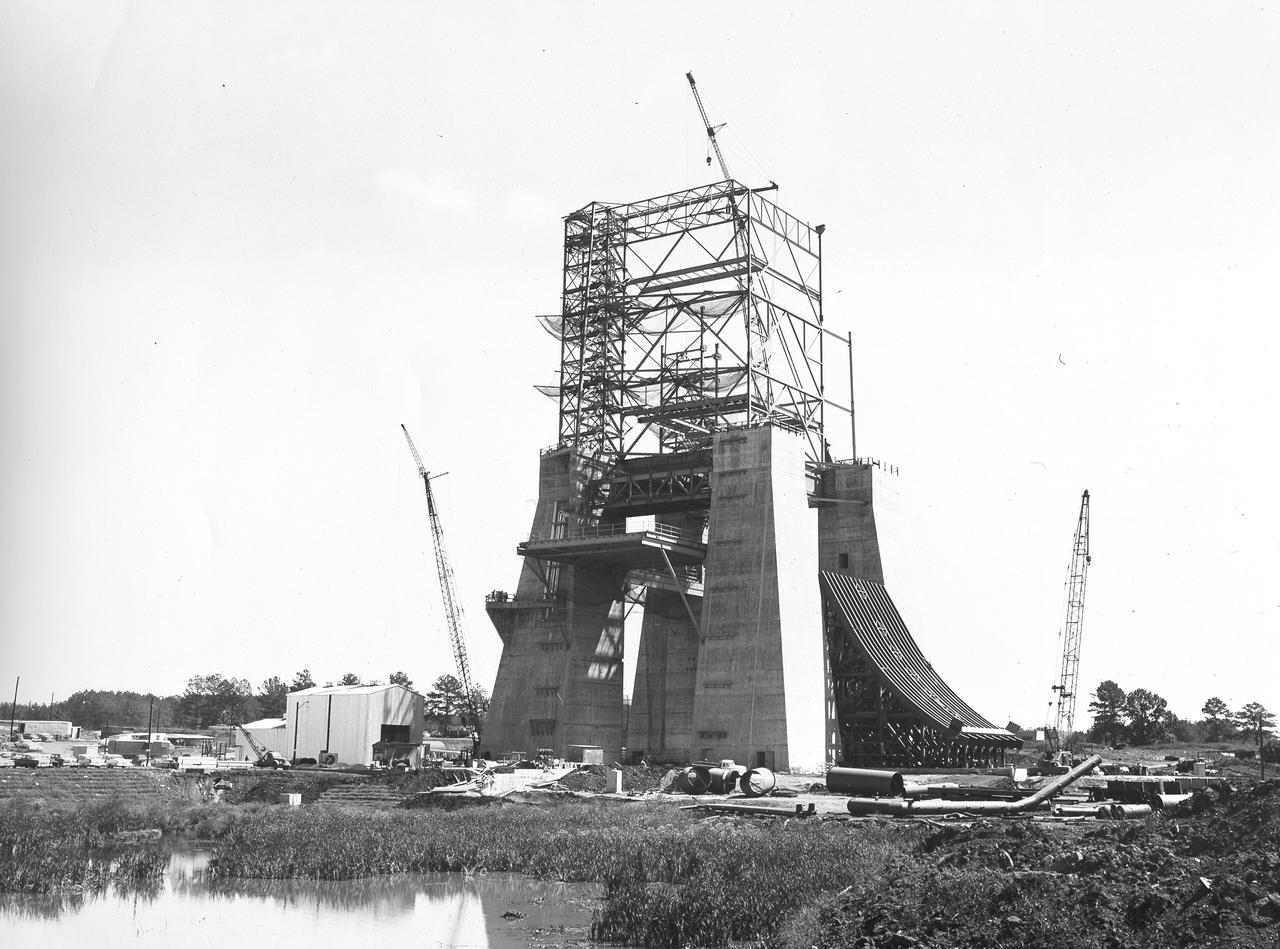 At its founding, the Marshall Space Flight Center (MSFC) inherited the Army’s Jupiter and Redstone test stands, but much larger facilities were needed for the giant stages of the Saturn V. From 1960 to 1964, the existing stands were remodeled and a sizable new test area was developed. The new comprehensive test complex for propulsion and structural dynamics was unique within the nation and the free world, and they remain so today because they were constructed with foresight to meet the future as well as on going needs. Construction of the S-IC Static test stand complex began in 1961 in the west test area of MSFC, and was completed in 1964. The S-IC static test stand was designed to develop and test the 138-ft long and 33-ft diameter Saturn V S-IC first stage, or booster stage, weighing in at 280,000 pounds. Required to hold down the brute force of a 7,500,000-pound thrust produced by 5 F-1 engines, the S-IC static test stand was designed and constructed with the strength of hundreds of tons of steel and 12,000,000 pounds of cement, planted down to bedrock 40 feet below ground level. The foundation walls, constructed with concrete and steel, are 4 feet thick. The base structure consists of four towers with 40-foot-thick walls extending upward 144 feet above ground level. The structure was topped by a crane with a 135-foot boom. With the boom in the upright position, the stand was given an overall height of 405 feet, placing it among the highest structures in Alabama at the time. In addition to the stand itself, related facilities were constructed during this time. Built to the northeast of the stand was a newly constructed Pump House. Its function was to provide water to the stand to prevent melting damage during testing. The water was sprayed through small holes in the stand’s 1900 ton flame deflector at the rate of 320,000 gallons per minute. In this photo of the S-IC test stand, taken September 25, 1963, the flame deflector can be seen rotated to the outside on the right. The deflector was assembled on tracks for mobility. 