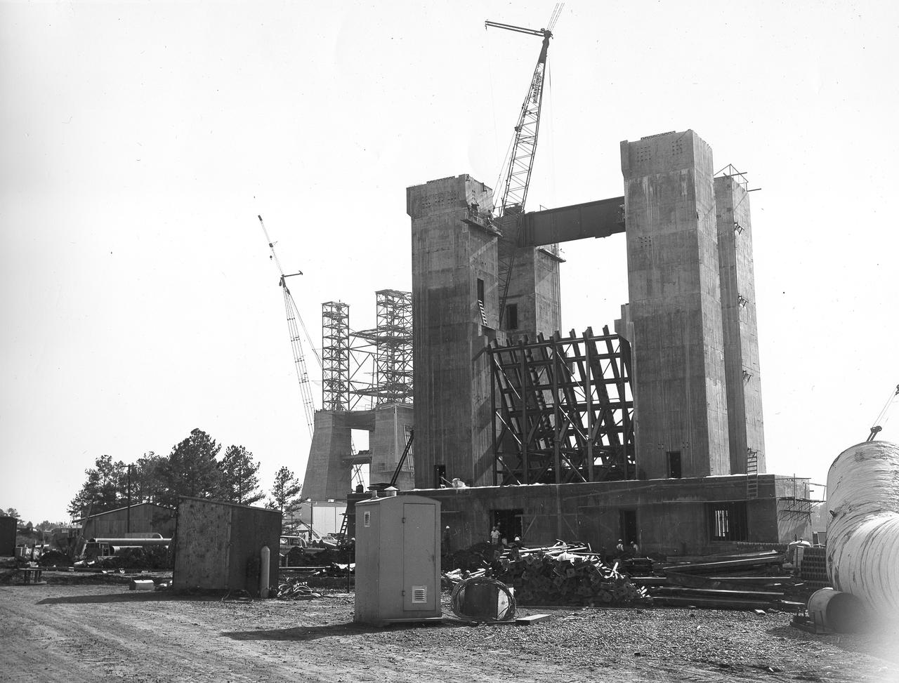 At its founding, the Marshall Space Flight Center (MSFC) inherited the Army’s Jupiter and Redstone test stands, but much larger facilities were needed for the giant stages of the Saturn V. From 1960 to 1964, the existing stands were remodeled and a sizable new test area was developed. The new comprehensive test complex for propulsion and structural dynamics was unique within the nation and the free world, and they remain so today because they were constructed with foresight to meet the future as well as on going needs. Construction of the S-IC Static test stand complex began in 1961 in the west test area of MSFC, and was completed in 1964. The S-IC static test stand was designed to develop and test the 138-ft long and 33-ft diameter Saturn V S-IC first stage, or booster stage, weighing in at 280,000 pounds. Required to hold down the brute force of a 7,500,000-pound thrust produced by 5 F-1 engines, the S-IC static test stand was designed and constructed with the strength of hundreds of tons of steel and 12,000,000 pounds of cement, planted down to bedrock 40 feet below ground level. The foundation walls, constructed with concrete and steel, are 4 feet thick. The base structure consists of four towers with 40-foot-thick walls extending upward 144 feet above ground level. The structure was topped by a crane with a 135-foot boom. With the boom in the upright position, the stand was given an overall height of 405 feet, placing it among the highest structures in Alabama at the time. In addition to the stand itself, related facilities were constructed during this time. North of the massive S-IC test stand, the F-1 Engine test stand was built. Designed to assist in the development of the F-1 Engine, the F-1 test stand is a vertical engine firing test stand, 239 feet in elevation and 4,600 square feet in area at the base. Capability was provided for static firing of 1.5 million pounds of thrust using liquid oxygen and kerosene. Like the S-IC stand, the foundation of the F-1 stand is keyed into the bedrock approximately 40 feet below grade. This photo depicts the construction of the F-1 test stand as of September 30, 1963. 