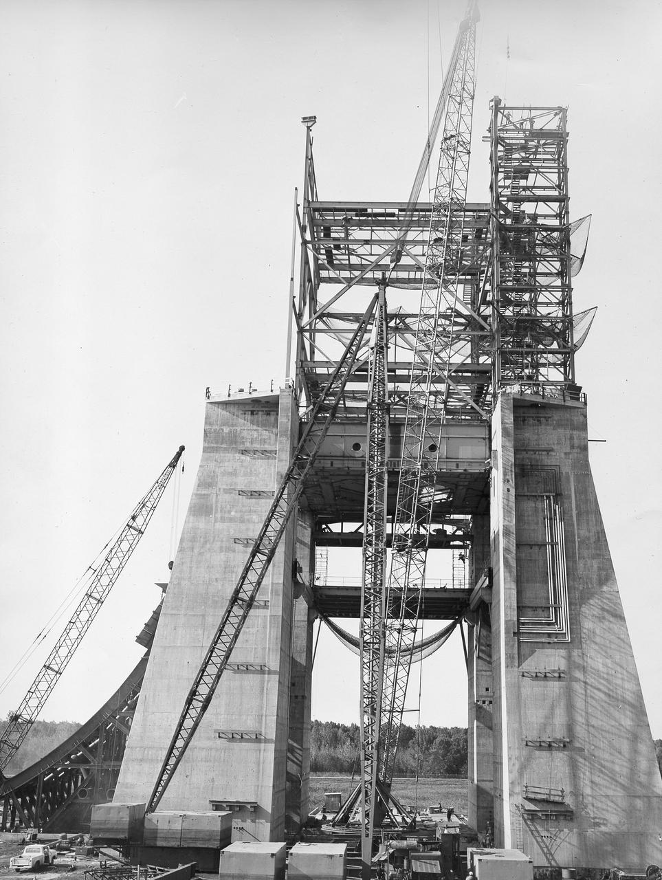 At its founding, the Marshall Space Flight Center (MSFC) inherited the Army’s Jupiter and Redstone test stands, but much larger facilities were needed for the giant stages of the Saturn V. From 1960 to 1964, the existing stands were remodeled and a sizable new test area was developed. The new comprehensive test complex for propulsion and structural dynamics was unique within the nation and the free world, and they remain so today because they were constructed with foresight to meet the future as well as on going needs. Construction of the S-IC Static test stand complex began in 1961 in the west test area of MSFC, and was completed in 1964. The S-IC static test stand was designed to develop and test the 138-ft long and 33-ft diameter Saturn V S-IC first stage, or booster stage, weighing in at 280,000 pounds. Required to hold down the brute force of a 7,500,000-pound thrust produced by 5 F-1 engines, the S-IC static test stand was designed and constructed with the strength of hundreds of tons of steel and 12,000,000 pounds of cement, planted down to bedrock 40 feet below ground level. The foundation walls, constructed with concrete and steel, are 4 feet thick. The base structure consists of four towers with 40-foot-thick walls extending upward 144 feet above ground level. The structure was topped by a crane with a 135-foot boom. With the boom in the upright position, the stand was given an overall height of 405 feet, placing it among the highest structures in Alabama at the time. In addition to the stand itself, related facilities were constructed during this time. Built to the northeast of the stand was a newly constructed Pump House. Its function was to provide water to the stand to prevent melting damage during testing. The water was sprayed through small holes in the stand’s 1900 ton flame deflector at the rate of 320,000 gallons per minute. In this photo of the S-IC test stand, taken September 25, 1963, the flame deflector can be seen rotated to the outside on the left. The deflector was assembled on tracks for mobility. 