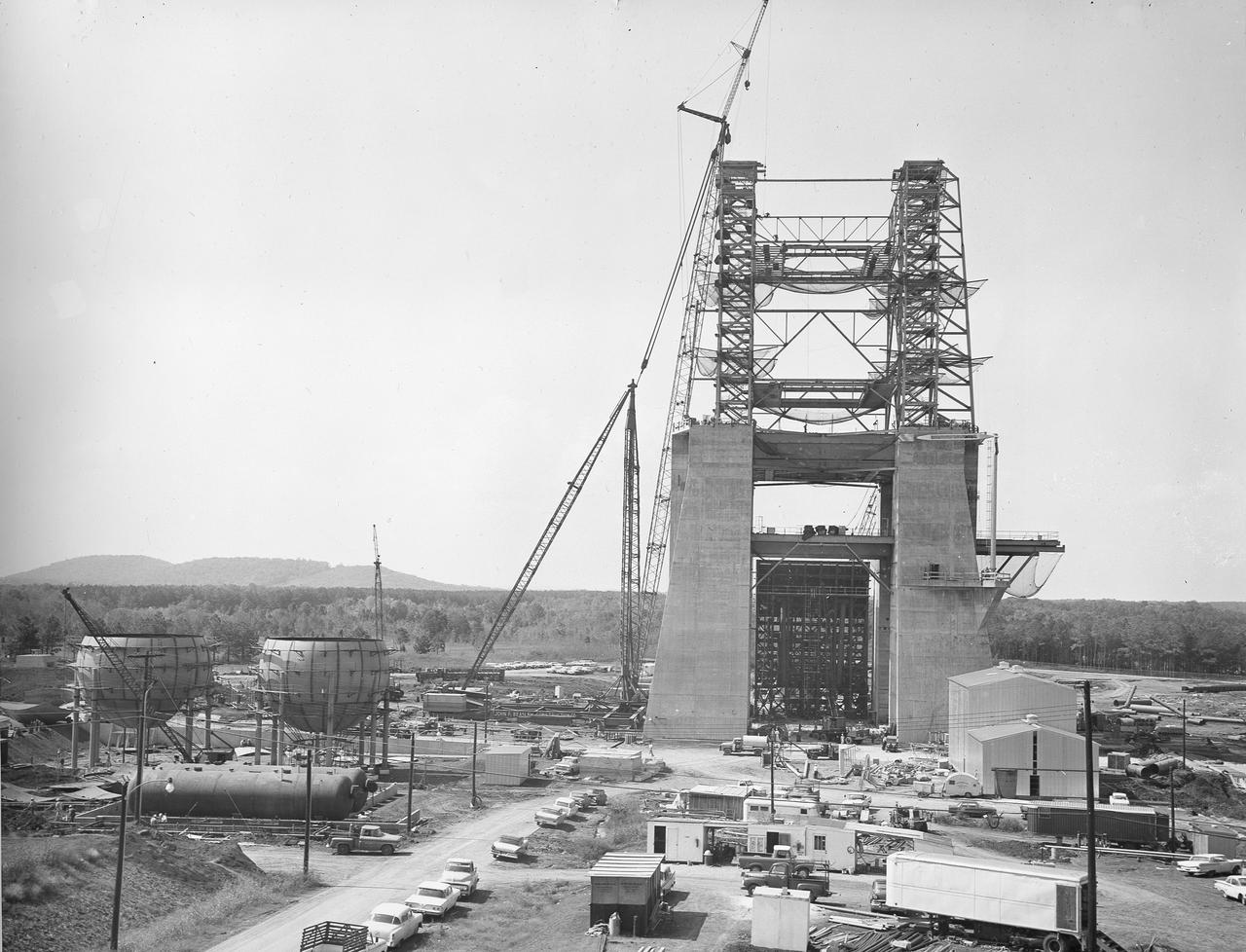 At its founding, the Marshall Space Flight Center (MSFC) inherited the Army’s Jupiter and Redstone test stands, but much larger facilities were needed for the giant stages of the Saturn V. From 1960 to 1964, the existing stands were remodeled and a sizable new test area was developed. The new comprehensive test complex for propulsion and structural dynamics was unique within the nation and the free world, and they remain so today because they were constructed with foresight to meet the future as well as on going needs. Construction of the S-IC Static test stand complex began in 1961 in the west test area of MSFC, and was completed in 1964. The S-IC static test stand was designed to develop and test the 138-ft long and 33-ft diameter Saturn V S-IC first stage, or booster stage, weighing in at 280,000 pounds. Required to hold down the brute force of a 7,500,000-pound thrust produced by 5 F-1 engines, the S-IC static test stand was designed and constructed with the strength of hundreds of tons of steel and 12,000,000 pounds of cement, planted down to bedrock 40 feet below ground level. The foundation walls, constructed with concrete and steel, are 4 feet thick. The base structure consists of four towers with 40-foot-thick walls extending upward 144 feet above ground level. The structure was topped by a crane with a 135-foot boom. With the boom in the upright position, the stand was given an overall height of 405 feet, placing it among the highest structures in Alabama at the time. In addition to the stand itself, related facilities were constructed during this time. Built to the northeast of the stand was a newly constructed Pump House. Its function was to provide water to the stand to prevent melting damage during testing. The water was sprayed through small holes in the stand’s 1900 ton flame deflector at the rate of 320,000 gallons per minute. In this photo of the S-IC test stand, taken October 2, 1963, the flame deflector can be seen in the bottom center portion of the stand. The deflector was assembled on tracks for mobility. To the left of the stand are two spherical hydrogen storage tanks.
