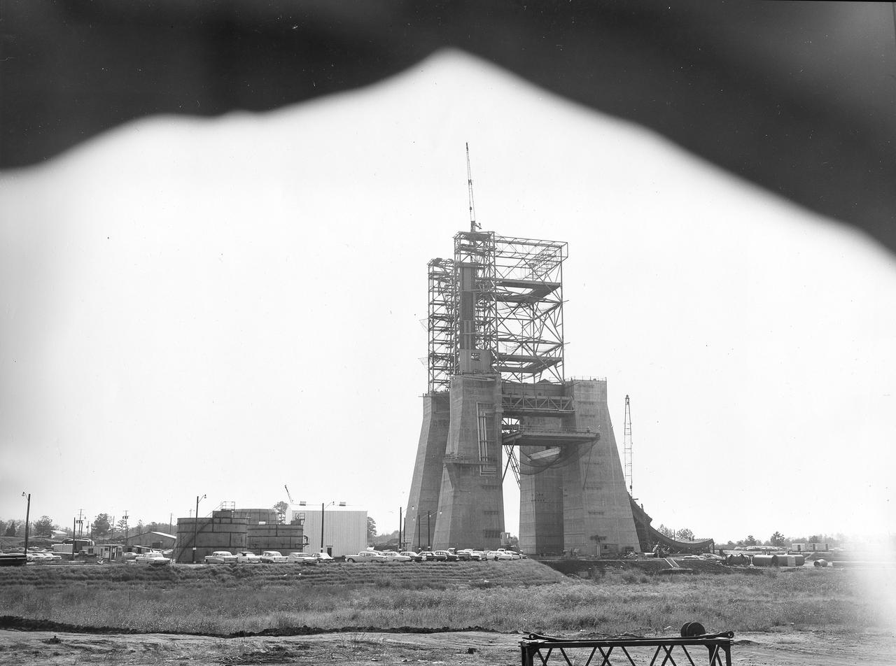At its founding, the Marshall Space Flight Center (MSFC) inherited the Army’s Jupiter and Redstone test stands, but much larger facilities were needed for the giant stages of the Saturn V. From 1960 to 1964, the existing stands were remodeled and a sizable new test area was developed. The new comprehensive test complex for propulsion and structural dynamics was unique within the nation and the free world, and they remain so today because they were constructed with foresight to meet the future as well as on going needs. Construction of the S-IC Static test stand complex began in 1961 in the west test area of MSFC, and was completed in 1964. The S-IC static test stand was designed to develop and test the 138-ft long and 33-ft diameter Saturn V S-IC first stage, or booster stage, weighing in at 280,000 pounds. Required to hold down the brute force of a 7,500,000-pound thrust produced by 5 F-1 engines, the S-IC static test stand was designed and constructed with the strength of hundreds of tons of steel and 12,000,000 pounds of cement, planted down to bedrock 40 feet below ground level. The foundation walls, constructed with concrete and steel, are 4 feet thick. The base structure consists of four towers with 40-foot-thick walls extending upward 144 feet above ground level. The structure was topped by a crane with a 135-foot boom. With the boom in the upright position, the stand was given an overall height of 405 feet, placing it among the highest structures in Alabama at the time. This photo shows the progress of the S-IC test stand as of October 10, 1963. Kerosene storage tanks can be seen to the left.