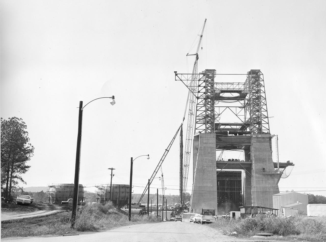 At its founding, the Marshall Space Flight Center (MSFC) inherited the Army’s Jupiter and Redstone test stands, but much larger facilities were needed for the giant stages of the Saturn V. From 1960 to 1964, the existing stands were remodeled and a sizable new test area was developed. The new comprehensive test complex for propulsion and structural dynamics was unique within the nation and the free world, and they remain so today because they were constructed with foresight to meet the future as well as on going needs. Construction of the S-IC Static test stand complex began in 1961 in the west test area of MSFC, and was completed in 1964. The S-IC static test stand was designed to develop and test the 138-ft long and 33-ft diameter Saturn V S-IC first stage, or booster stage, weighing in at 280,000 pounds. Required to hold down the brute force of a 7,500,000-pound thrust produced by 5 F-1 engines, the S-IC static test stand was designed and constructed with the strength of hundreds of tons of steel and 12,000,000 pounds of cement, planted down to bedrock 40 feet below ground level. The foundation walls, constructed with concrete and steel, are 4 feet thick. The base structure consists of four towers with 40-foot-thick walls extending upward 144 feet above ground level. The structure was topped by a crane with a 135-foot boom. With the boom in the upright position, the stand was given an overall height of 405 feet, placing it among the highest structures in Alabama at the time. This photo shows the progress of the S-IC test stand as of October 10, 1963. Spherical liquid hydrogen tanks can be seen to the left.