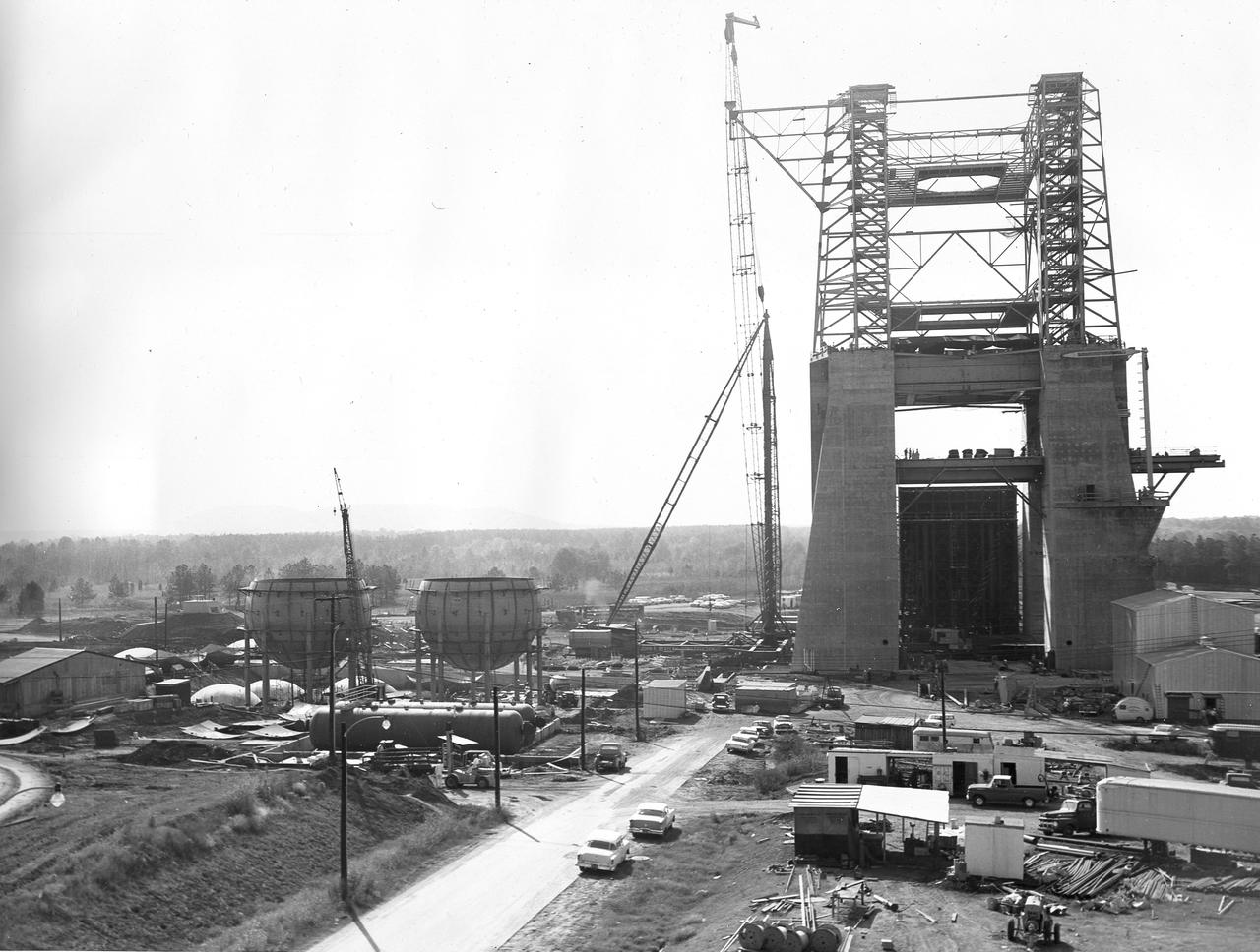 At its founding, the Marshall Space Flight Center (MSFC) inherited the Army’s Jupiter and Redstone test stands, but much larger facilities were needed for the giant stages of the Saturn V. From 1960 to 1964, the existing stands were remodeled and a sizable new test area was developed. The new comprehensive test complex for propulsion and structural dynamics was unique within the nation and the free world, and they remain so today because they were constructed with foresight to meet the future as well as on going needs. Construction of the S-IC Static test stand complex began in 1961 in the west test area of MSFC, and was completed in 1964. The S-IC static test stand was designed to develop and test the 138-ft long and 33-ft diameter Saturn V S-IC first stage, or booster stage, weighing in at 280,000 pounds. Required to hold down the brute force of a 7,500,000-pound thrust produced by 5 F-1 engines, the S-IC static test stand was designed and constructed with the strength of hundreds of tons of steel and 12,000,000 pounds of cement, planted down to bedrock 40 feet below ground level. The foundation walls, constructed with concrete and steel, are 4 feet thick. The base structure consists of four towers with 40-foot-thick walls extending upward 144 feet above ground level. The structure was topped by a crane with a 135-foot boom. With the boom in the upright position, the stand was given an overall height of 405 feet, placing it among the highest structures in Alabama at the time. This photo shows the progress of the S-IC test stand as of October 22, 1963. Spherical liquid hydrogen tanks can be seen to the left. Just to the lower front of those are the cylindrical liquid oxygen (LOX) tanks.