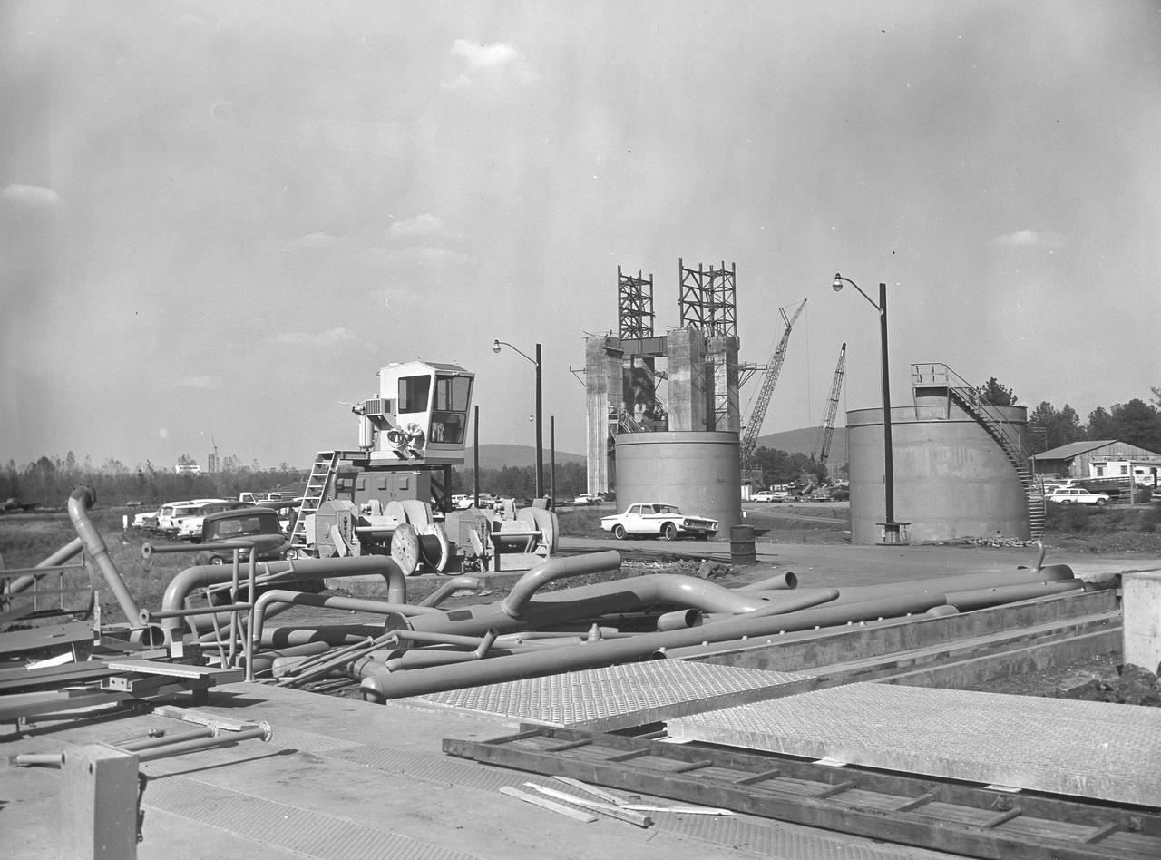 At its founding, the Marshall Space Flight Center (MSFC) inherited the Army’s Jupiter and Redstone test stands, but much larger facilities were needed for the giant stages of the Saturn V. From 1960 to 1964, the existing stands were remodeled and a sizable new test area was developed. The new comprehensive test complex for propulsion and structural dynamics was unique within the nation and the free world, and they remain so today because they were constructed with foresight to meet the future as well as on going needs. Construction of the S-IC Static test stand complex began in 1961 in the west test area of MSFC, and was completed in 1964. The S-IC static test stand was designed to develop and test the 138-ft long and 33-ft diameter Saturn V S-IC first stage, or booster stage, weighing in at 280,000 pounds. Required to hold down the brute force of a 7,500,000-pound thrust produced by 5 F-1 engines, the S-IC static test stand was designed and constructed with the strength of hundreds of tons of steel and 12,000,000 pounds of cement, planted down to bedrock 40 feet below ground level. The foundation walls, constructed with concrete and steel, are 4 feet thick. The base structure consists of four towers with 40-foot-thick walls extending upward 144 feet above ground level. The structure was topped by a crane with a 135-foot boom. With the boom in the upright position, the stand was given an overall height of 405 feet, placing it among the highest structures in Alabama at the time. In addition to the stand itself, related facilities were constructed during this time. Northeast of the massive S-IC test stand, the F-1 Engine test stand was built. The F-1 test stand is a vertical engine firing test stand, 239 feet in elevation and 4,600 square feet in area at the base, and was designed to assist in the development of the F-1 Engine. Capability was provided for static firing of 1.5 million pounds of thrust using liquid oxygen and kerosene. Like the S-IC stand, the foundation of the F-1 stand is keyed into the bedrock approximately 40 feet below grade. This photo depicts the fuel tanks that housed kerosene and just beyond those is the F-1 test stand.