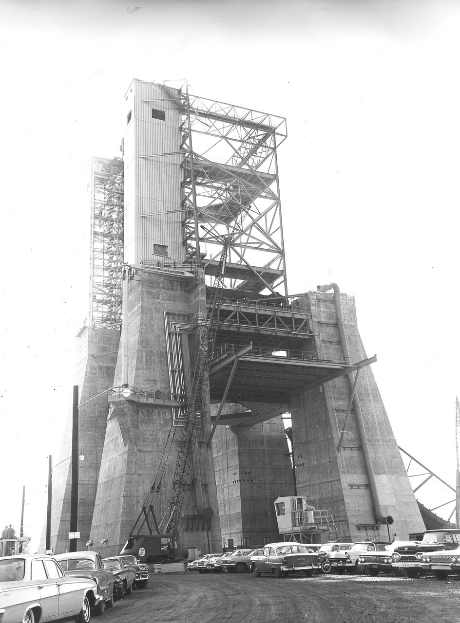 At its founding, the Marshall Space Flight Center (MSFC) inherited the Army’s Jupiter and Redstone test stands, but much larger facilities were needed for the giant stages of the Saturn V. From 1960 to 1964, the existing stands were remodeled and a sizable new test area was developed. The new comprehensive test complex for propulsion and structural dynamics was unique within the nation and the free world, and they remain so today because they were constructed with foresight to meet the future as well as on going needs. Construction of the S-IC Static test stand complex began in 1961 in the west test area of MSFC, and was completed in 1964. The S-IC static test stand was designed to develop and test the 138-ft long and 33-ft diameter Saturn V S-IC first stage, or booster stage, weighing in at 280,000 pounds. Required to hold down the brute force of a 7,500,000-pound thrust produced by 5 F-1 engines, the S-IC static test stand was designed and constructed with the strength of hundreds of tons of steel and 12,000,000 pounds of cement, planted down to bedrock 40 feet below ground level. The foundation walls, constructed with concrete and steel, are 4 feet thick. The base structure consists of four towers with 40-foot-thick walls extending upward 144 feet above ground level. The structure was topped by a crane with a 135-foot boom. With the boom in the upright position, the stand was given an overall height of 405 feet, placing it among the highest structures in Alabama at the time. This photo shows the progress of the S-IC test stand as of November 20, 1963. 