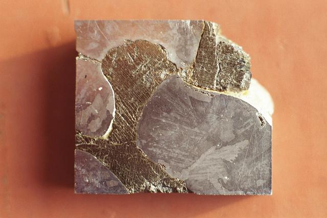 This is a macro photograph of an etched surface of the Mundrabilla meteorite, a small piece of the approximately 3.9 billion-year-old meteorite that was first discovered in Western Australia in 1911. Two more giant chunks, together weighing about 17 tons, were found in 1966. Researchers can learn much from this natural crystal growth experiment since it has spent several hundred million years cooling, and would be impossible to emulate in a lab. This single slice, taken from a 6 ton piece recovered in 1966, measures only 2 square inches. The macro photograph shows a metallic iron-nickel alloy phase of kamcite (38% Ni) and taenite (6% Ni) at bottom right, bottom left, and top left.  The darker material is an iron sulfide (FeS or troilite) with a parallel precipitates of duabreelite (iron chromium sulfide (FeCr2S4).