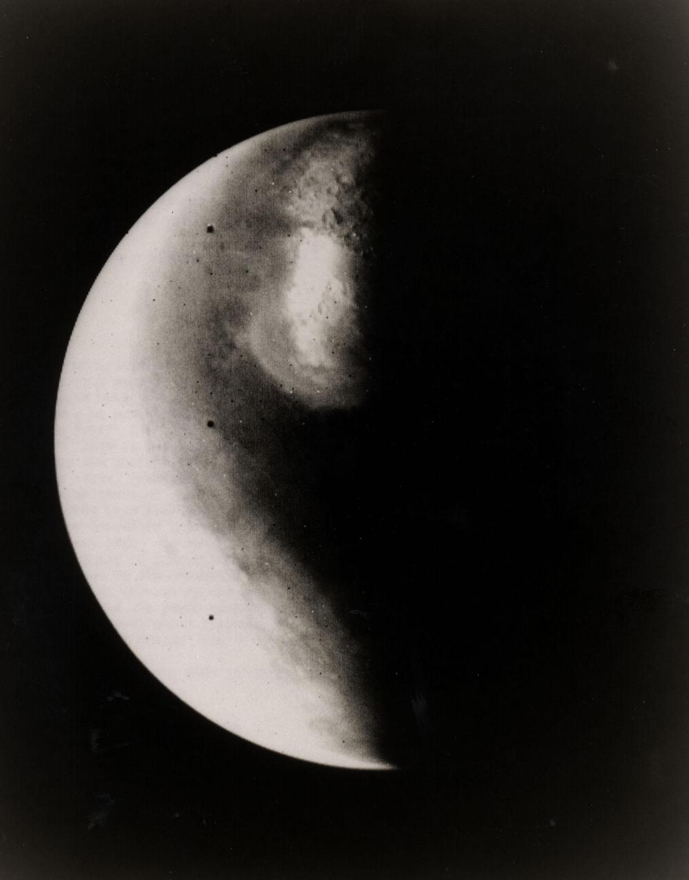 Half-Mars hangs in space 685,000 kilometers (425,000 miles) above Viking 1 as the spacecraft approaches the planet. This picture was taken at 6:40 p.m. PDT on June 16 by one of two telescope-equipped TV cameras aboard the Viking 1 Orbiter. A violet filter was used. Resolution is approximately 17 kilometers (10.5 miles) at center of disk. North is toward upper right and the south pole is in the dark to the lower left. We are looking at the morning side of the planet; that is, the planet is rotating from left to right. Toward the bottom of the image is a very bright irregular feature within a somewhat less bright circular feature. The circular feature is Hellas, a 2,000-kilometer(1,250-mile) diameter impact basin. Numerous craters are just visible within the frost-covered region. To the south of Hellas is another brighter area. This is probably an area of discontinuous frost cover around the south pole. The image is very bright toward the edge of the planet because of atmospheric scattering in the violet. Viking 1 will begin orbiting Mars on Saturday, June 19, with the landing planned for July 4. The Viking Project is managed by the NASA Langley Reseach Center, Hampton, Va.