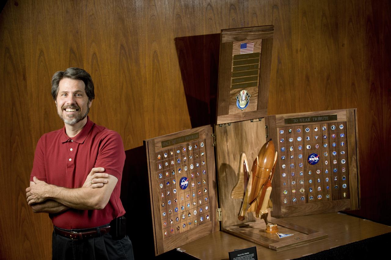 SCOTT PHILLIPS STANDS NEAR HIS LATEST WOODWORKING CREATION FEATURING A SHUTTLE MODEL AND ALL OF THE MISSION PINS FROM PREVIOUS LAUNCHES.