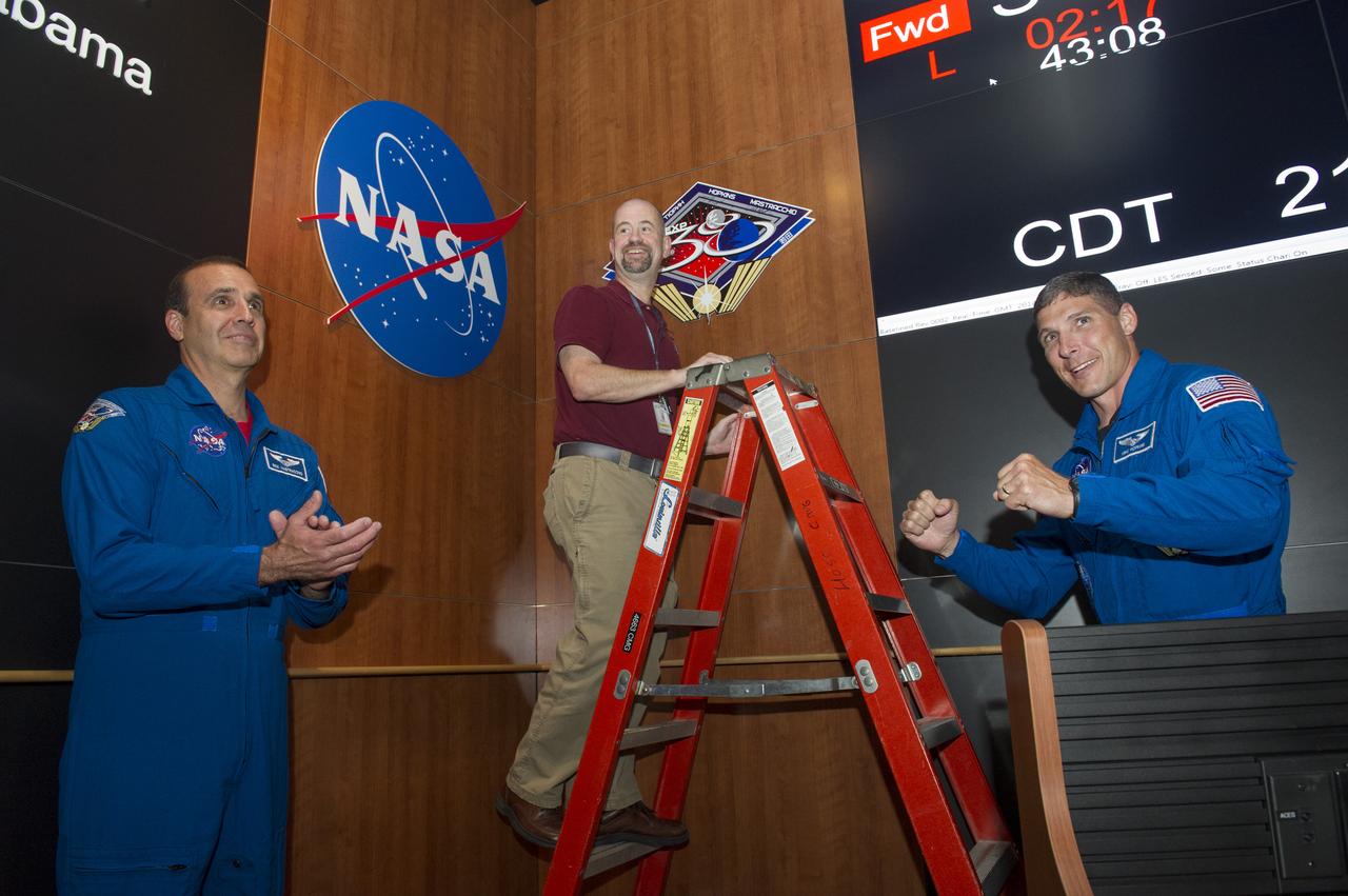 NASA ASTRONAUTS RICHARD MASTRACCHIO, LEFT, AND MICHAEL HOPKINS FLANK ED LITKENHOUS, A PAYLOAD ACTIVITY REQUIREMENTS COORDINATOR IN THE PAYLOAD OPERATIONS AND INTEGRATION CENTER (POIC), AS HE HANGS THE ASTRONAUTS' MISSION PLAQUE ON THE WALL OF THE POIC IN THE MARSHALL CENTER'S BUILDING 4663. THE PLAQUE JOINED THE LOGOS FROM PREVIOUS MISSIONS HANGING AROUND THE ROOM, RECOGNIZING THE COLLABORATION BETWEEN THE ORBITING ASTRONAUTS AND THE HUNTSVILLE-BASED TEAM