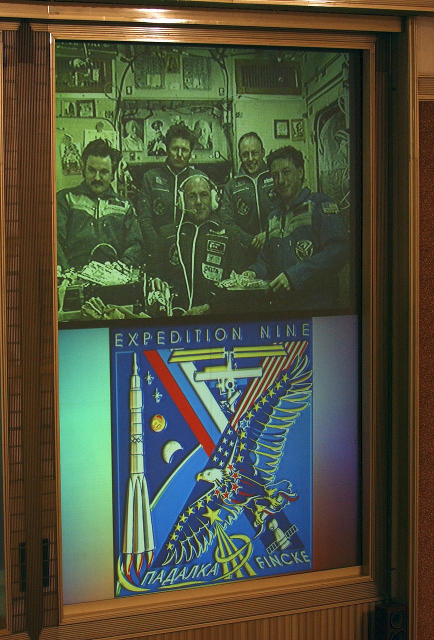 The Expedition 8 and 9 crews and European Space Agency astronaut Andre Kuipers of the Netherlands are viewed on the front screen of the Flight Control Room at the Russian Mission Control Center outside Moscow, Wednesday, April 21, 2004, in a televised welcoming ceremony following their docking to the International Space Station in a Russian Soyuz spacecraft. Under the televised view of the two crews is the insignia of the Expedition 9 crew, consisting of commander Gennady Padalka and Flight Engineer and NASA International Space Station Science Officer Michael Fincke, who will spend six months on the Station. Photo Credit: (NASA/Bill Ingalls)