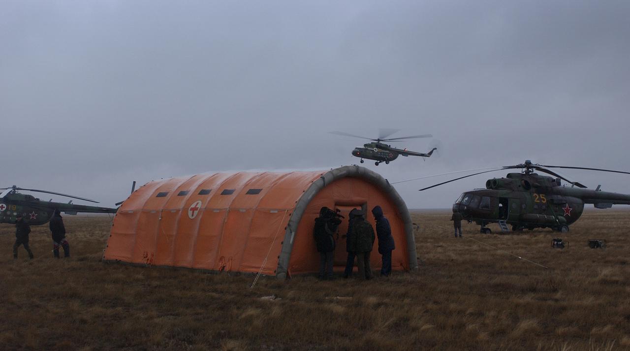 An inflatable medical tent stands in the foreground of the Expedition 9 landing site, while an incoming Russian Search and Rescue helicopter lands. The Soyuz capsule, which carried Expedition 9 Flight Engineer Michael Fincke, Commander Gennady Padalka and Russian Space Forces cosmonaut Yuri Shargin landed approximately 85 kilometers northeast of Arkalyk in northern Kazakhstan, Sunday, October 24, 2004.  Photo Credit: (NASA/Bill Ingalls)