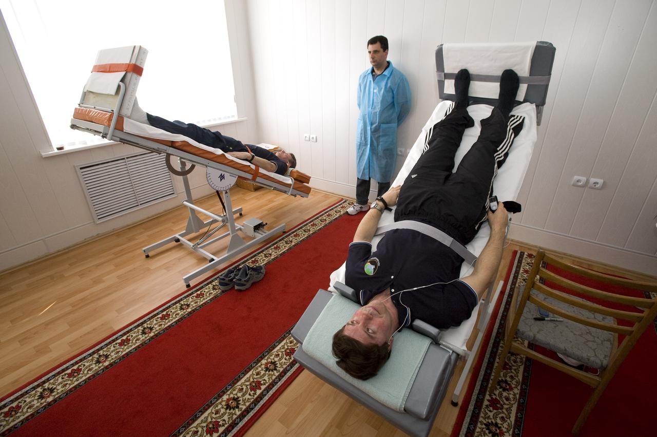 Expedition 19 Commander Gennady I. Padalka, left, and Flight Engineer Michael R. Barratt listen to their mp3 players as a medical doctor looks on during the tilt table training at the Cosmonaut Hotel, Saturday, March 21, 2009 in Baikonur, Kazakhstan.(Photo Credit: NASA/Bill Ingalls)