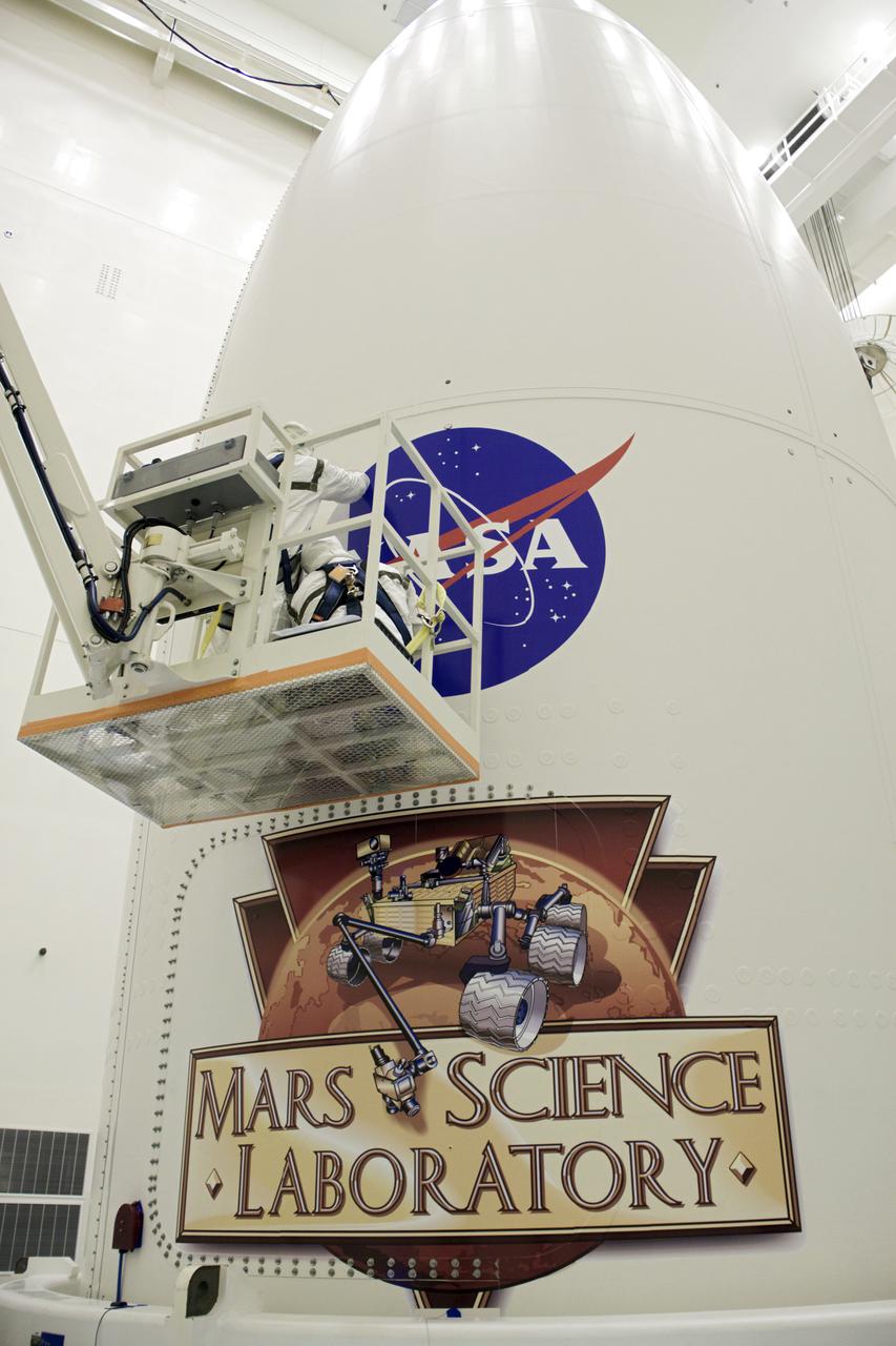 CAPE CANAVERAL, Fla. -- In the Payload Hazardous Servicing Facility at NASA's Kennedy Space Center in Florida, technicians use a lift to reach the upper region of an Atlas V rocket's payload fairing, where the NASA logo is being installed. Below is the Mars Science Laboratory (MSL) mission logo. Tucked inside the fairing is the MSL spacecraft, including a compact car-sized rover, Curiosity, which has 10 science instruments designed to search for evidence on whether Mars has had environments favorable to microbial life, including the chemical ingredients for life. The unique rover will use a laser to look inside rocks and release its gasses so that the rover’s spectrometer can analyze and send the data back to Earth. Launch of MSL aboard a United Launch Alliance Atlas V rocket is planned for Nov. 25 from Space Launch Complex-41 on Cape Canaveral Air Force Station. For more information, visit http:__www.nasa.gov_msl. Photo credit: NASA_Jim Grossmann