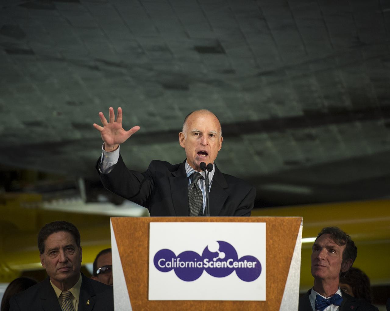 California Governor Jerry Brown speaks from a podium underneath the space shuttle Endeavour during the grand opening ceremony for the California Science center's Samuel Oschin Space Shuttle Endeavour Display Pavilion, Tuesday, Oct. 30, 2012, in Los Angeles.  Endeavour, built as a replacement for space shuttle Challenger, completed 25 missions, spent 299 days in orbit, and orbited Earth 4,671 times while traveling 122,883,151 miles. Photo Credit: (NASA/Bill Ingalls)