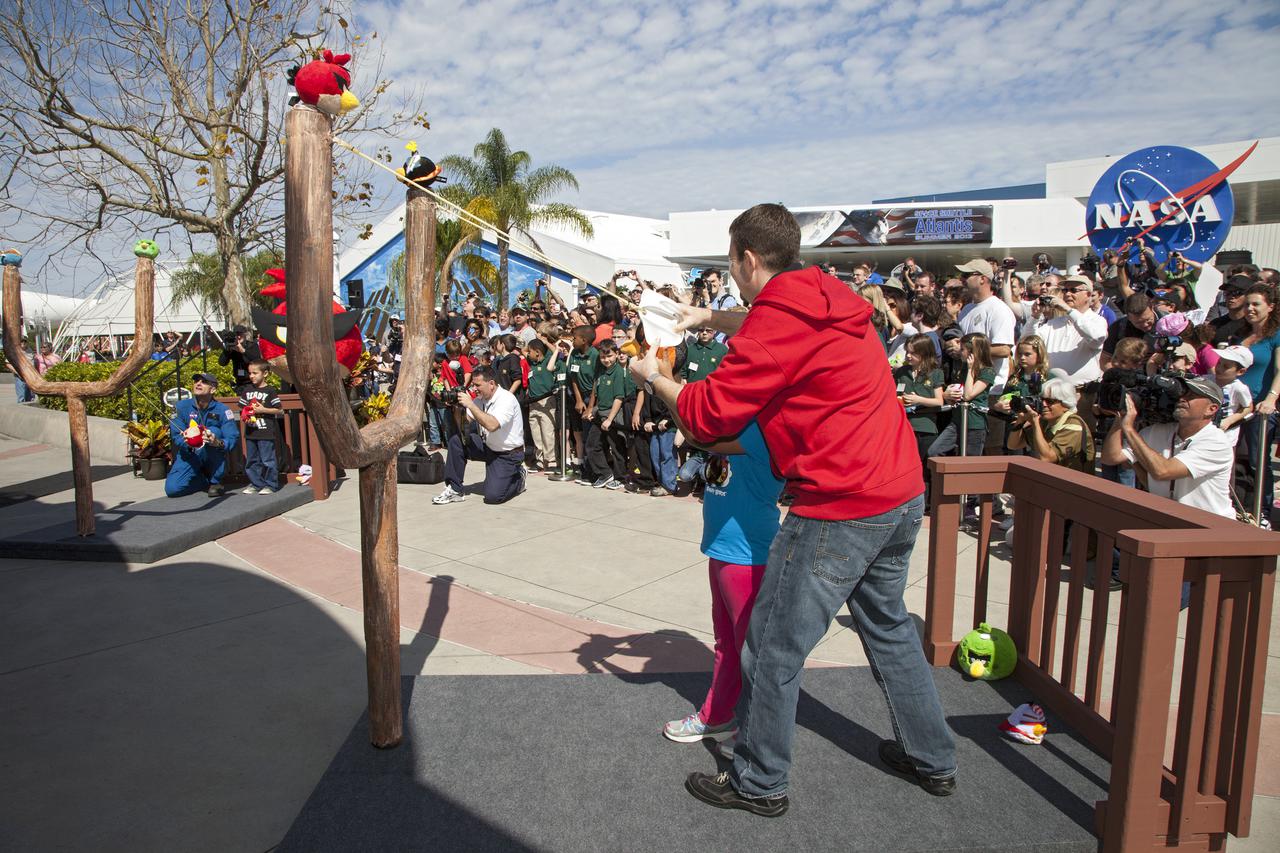 CAPE CANAVERAL, Fla. -- At NASA’s Kennedy Space Center Visitor Complex in Florida, Dan Mitchell, director of location-based entertainment for Rovio Entertainment, uses a giant slingshot to launch a plush Angry Bird character during the grand opening of the new Angry Birds Space Encounter. To Mitchell’s left is NASA astronaut Don Pettit.  Finland-based Rovio Entertainment, the creator of the Angry Birds Space game, partnered with Kennedy Space Center to bring the beloved characters to life. It is the first Angry Birds interactive exhibit in the United States designed for people of all ages. The 4,485-square-foot facility hosts the space adventures of the Angry Birds as they travel into an intergalactic wormhole, come face-to-face with Space Pigs and discover heroic superpowers. Six interactive stations are designed to encourage children in science, technology, engineering and mathematics fields of study. Photo credit: NASA_Kim Shiflett