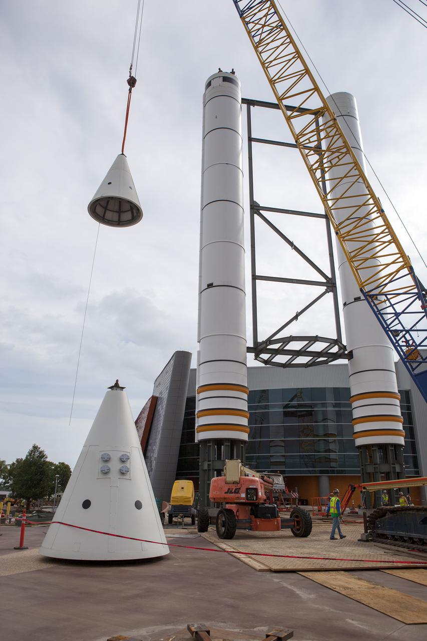 CAPE CANAVERAL, Fla. – A crane lifts a nose cone to top out one of a pair of replica space shuttle solid rocket boosters at the entry of the space shuttle Atlantis attraction under construction at the Kennedy Space Center Visitor Complex. Workers will use cranes to place the nose cones atop each of the model boosters. The building in the background houses the actual shuttle Atlantis. Photo credit: NASA_Dmitri Gerondidakis