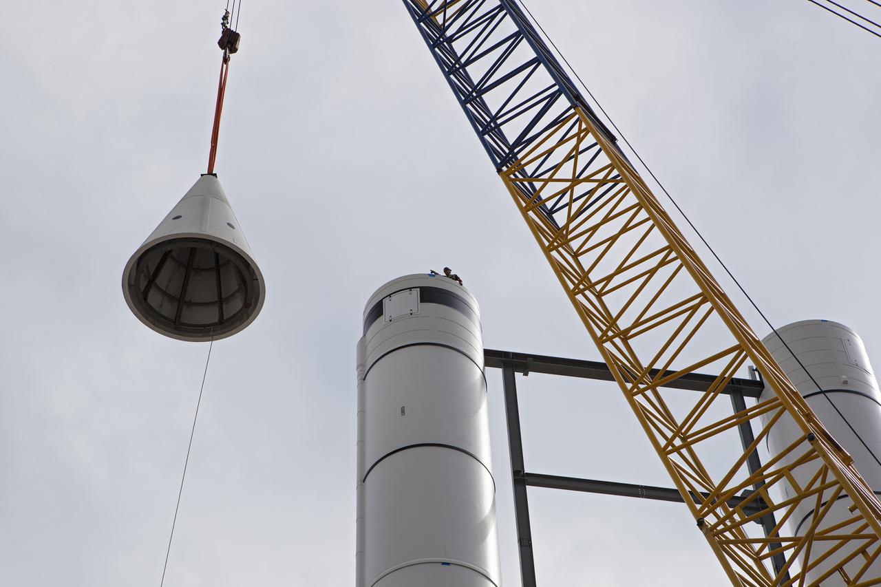 CAPE CANAVERAL, Fla. – A crane lifts a nose cone to top out one of a pair of replica space shuttle solid rocket boosters at the entry of the space shuttle Atlantis attraction under construction at the Kennedy Space Center Visitor Complex. Workers will use cranes to place the nose cones atop each of the model boosters. The building in the background houses the actual shuttle Atlantis. Photo credit: NASA_Dmitri Gerondidakis