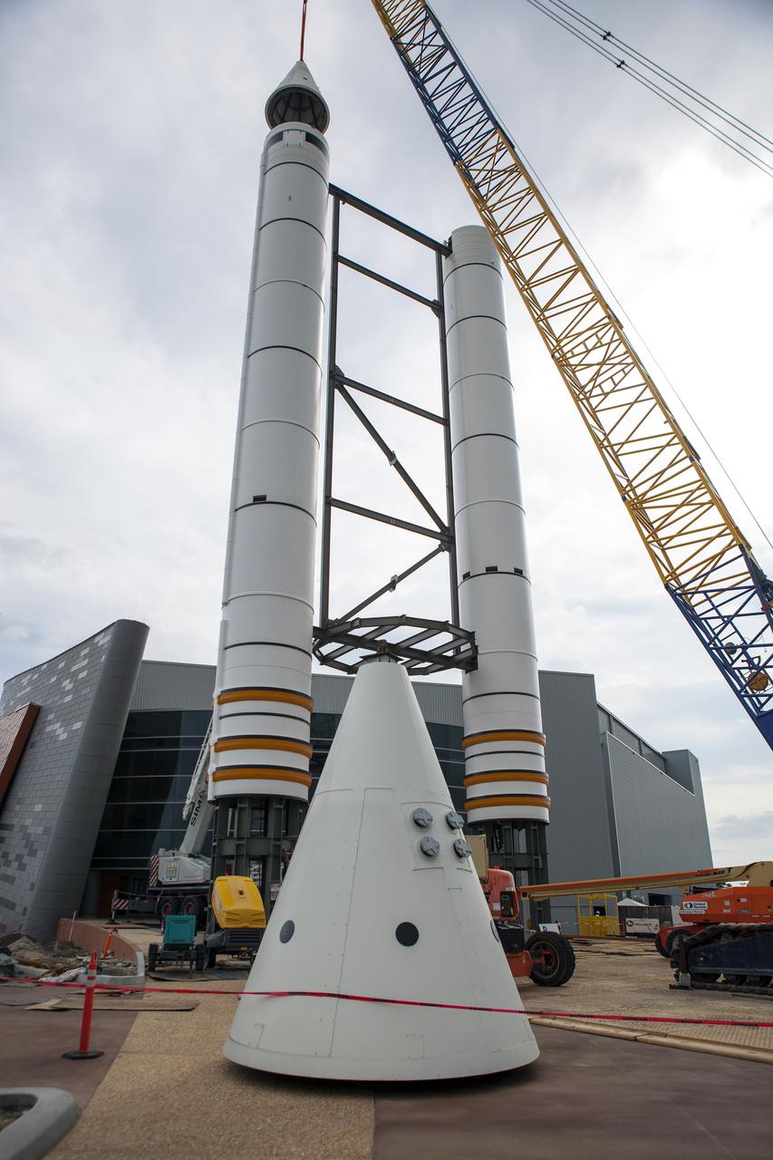 CAPE CANAVERAL, Fla. – A crane lifts a nose cone to top out one of a pair of replica space shuttle solid rocket boosters at the entry of the space shuttle Atlantis attraction under construction at the Kennedy Space Center Visitor Complex. Workers will use cranes to place the nose cones atop each of the model boosters. The building in the background houses the actual shuttle Atlantis. Photo credit: NASA_Dmitri Gerondidakis