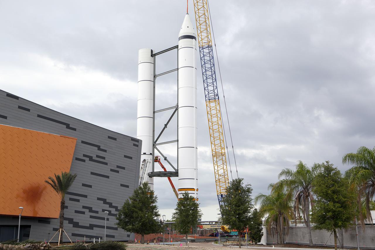 CAPE CANAVERAL, Fla. – A crane places a nose cone to top out one of a pair of replica space shuttle solid rocket boosters at the entry of the space shuttle Atlantis attraction under construction at the Kennedy Space Center Visitor Complex. Workers will use cranes to place the nose cones atop each of the model boosters. The building in the background houses the actual shuttle Atlantis. Photo credit: NASA_Dmitri Gerondidakis