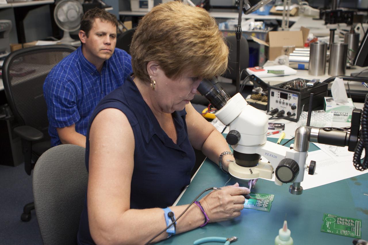 CAPE CANAVERAL, Fla. – Theresa Overcash works with elements of a prototype of the StangSat at Kennedy Space Center before final assembly as Shaun Daly looks on. The satellite is a small cube measuring 10 inches on all sides and will be launched on a rocket that will carry it on a suborbital mission in Mojave, Calif. Photo credit: NASA_Kim Shiflett