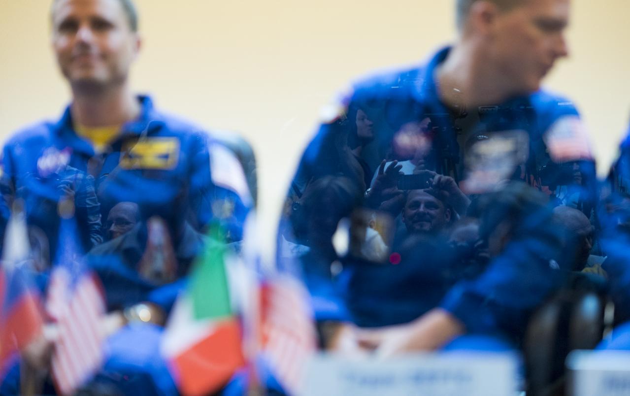 A man taking a picture with a cell phone is seen reflected in the glass separating the quarantined crew during a press conference on Tuesday, May 27, 2014, at the Cosmonaut Hotel in Baikonur, Kazakhstan. The mission to the International Space Station is set to launch May 29 from the Baikonur Cosmodrome. Photo Credit: (NASA/Joel Kowsky)