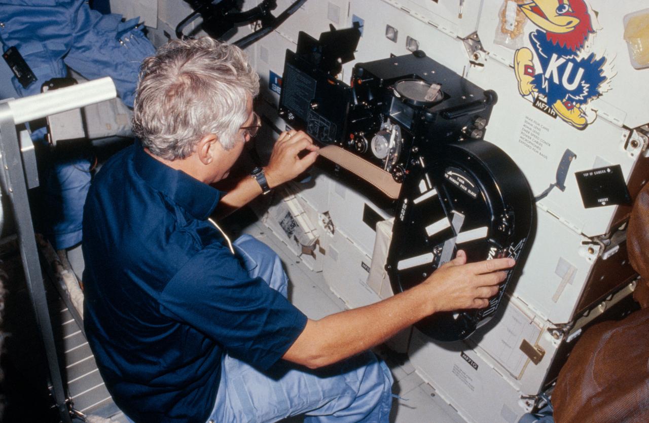 41D-11-004 (8 September 1984 --- View of Crew Commander Henry Hartsfield Jr. loading film into the IMAX camera during the 41-D mission. The camera is floating in front of the middeck lockers. Above it is a sticker of the University of Kansas mascott, the Jayhawk.