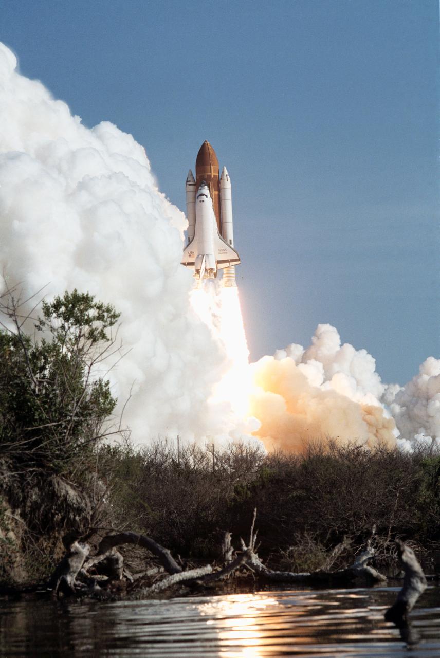View of the launch of the shuttle Discovery and the 41-D mission. The orbiter and its plume of smoke are photographed from across the river in the early stages of liftoff (3085,3087); The orbiter can be seen just clearing the pad, with a cloud of smoke billowing to the left of the frame. The orbiters two orbital maneuvering system (OMS) pods and external tank are visible along with the orbiter (3086).