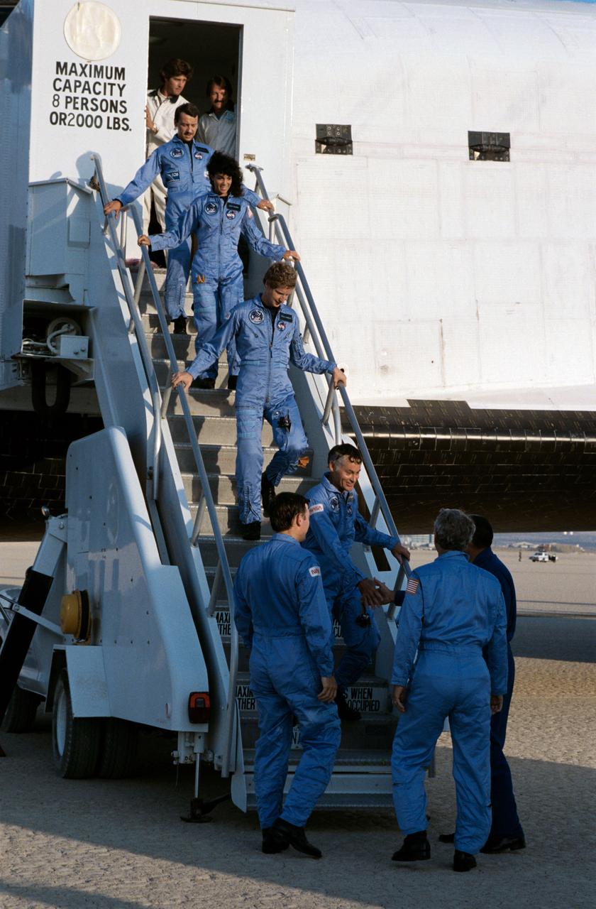 The crew of the STS 41-D mission exit the orbiter after landing at Edwards Air Force Base in California. Starting at the top of the ladder is Astronaut Charles D. Walker, payload specialist; Judith A. Resnik, mission specialist; Steven A. Hawley, mission specialist; and Richard M. Mike Mullane, mission specialist. Waiting at the bottom of the ramp are Astronaut Mike Coats (left), pilot and Henry W. Hartsfield, Jr. (center), crew commander.