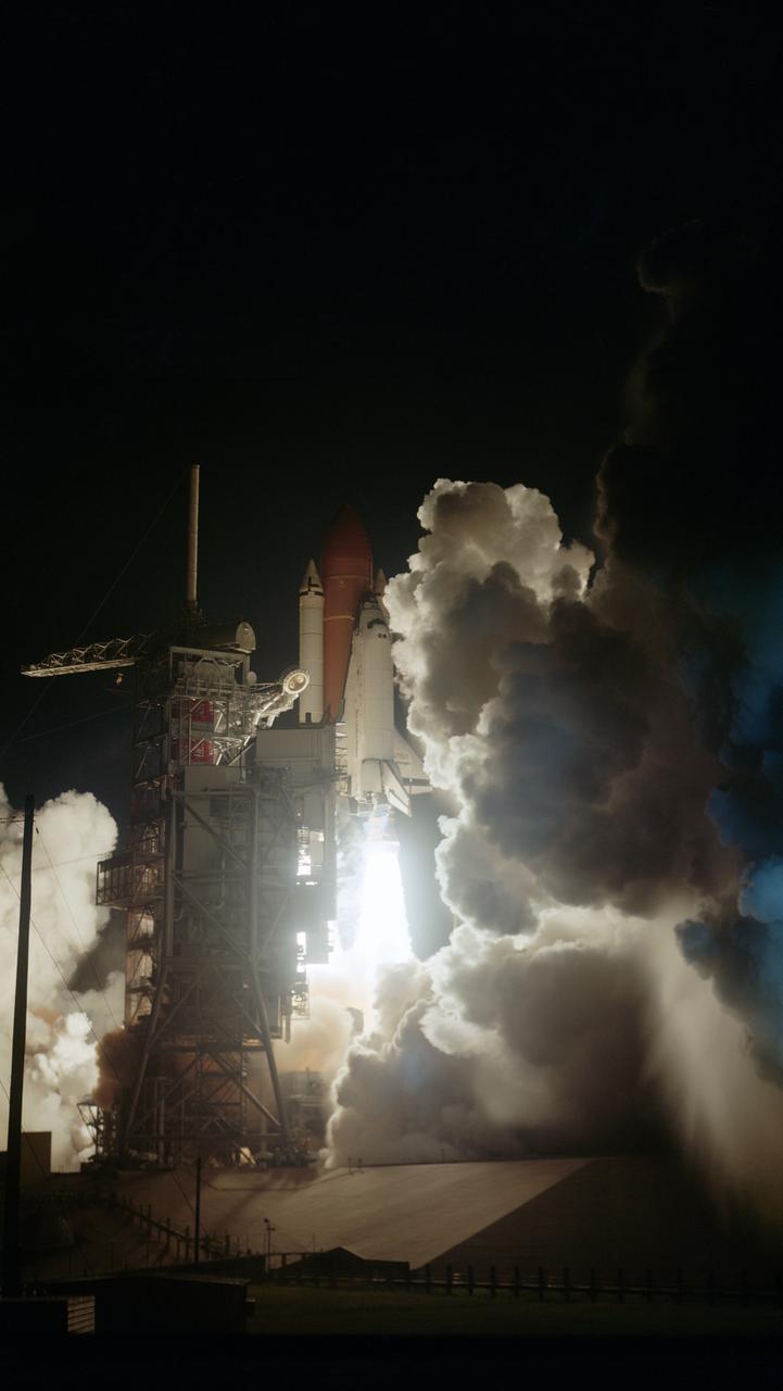 View of the early morning launch of STS 41-G Challenger. The dark launch complex is illuminated by spotlights as the orbiter begins its ascent from the pad. The light is reflected off the clouds of smoke from the orbiter's engines.