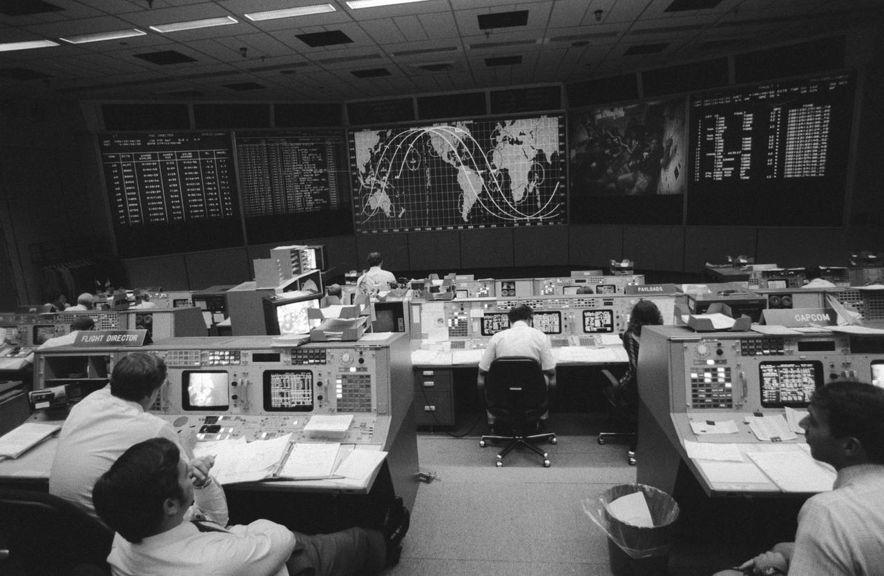 View from the back of the Mission Control Center (MCC). Visible are the Flight Directors console (left front), the CAPCOM console (right front) and the Payloads console. Some of the STS 41-G crew can be seen on a large screen at the front of the MCC along with a map tracking the progress of the orbiter.