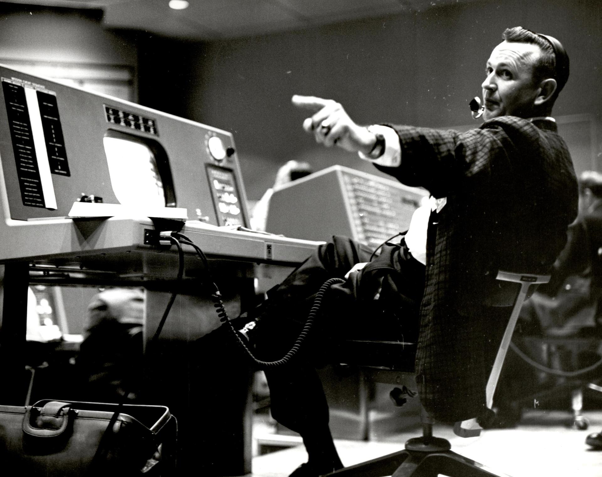 Chris Kraft sits at at a console in mission control pointing to his left