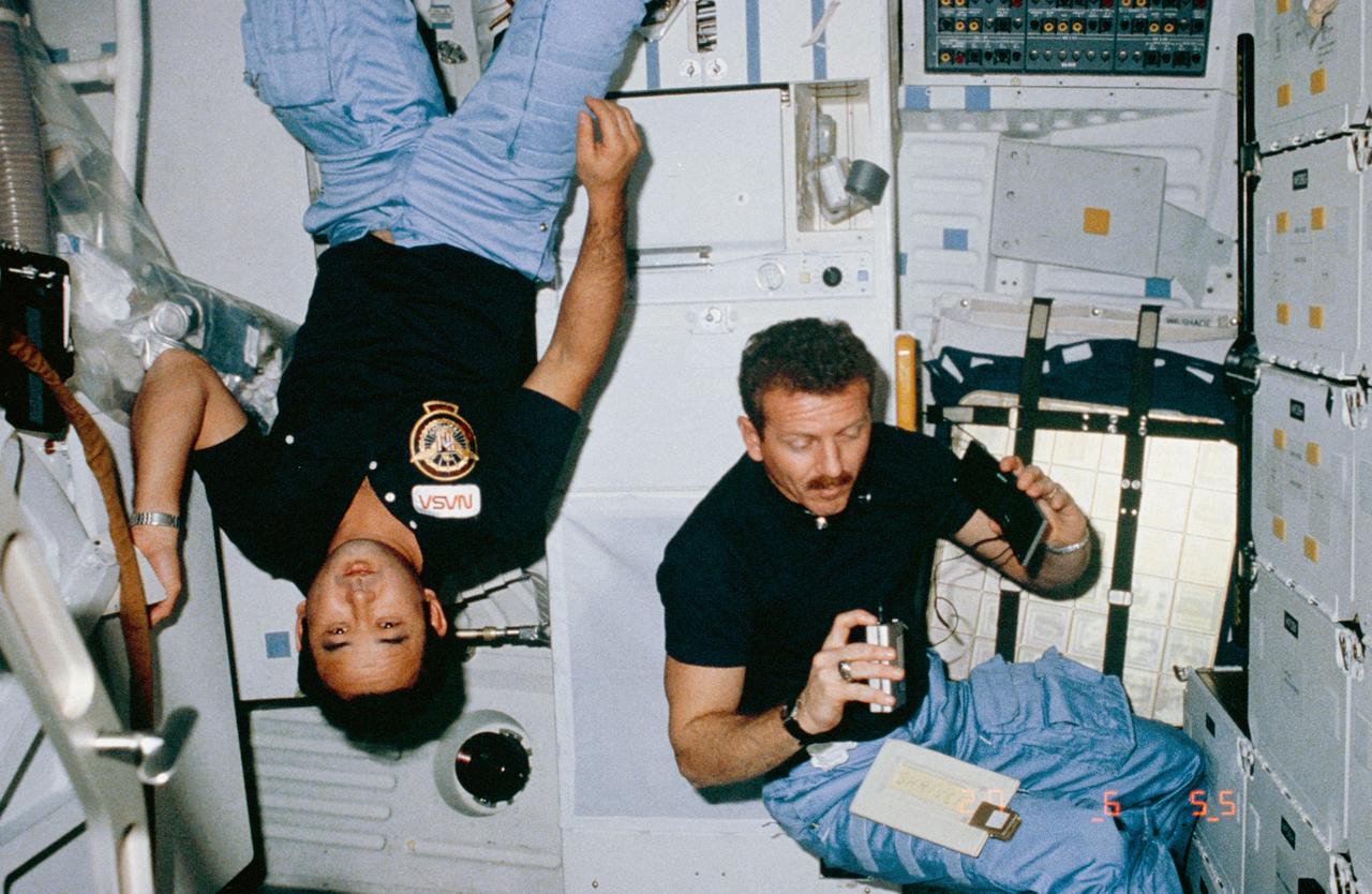 51C-08-017 (24-27 Jan 1985) --- Astronaut Ellison S. Onizuka, mission specialist, (left) turns 180 degrees near airlock hatch, while Loren J. Shriver, pilot, records verbal mission - pertinent notes. For orientation hold the picture with lockers on right.