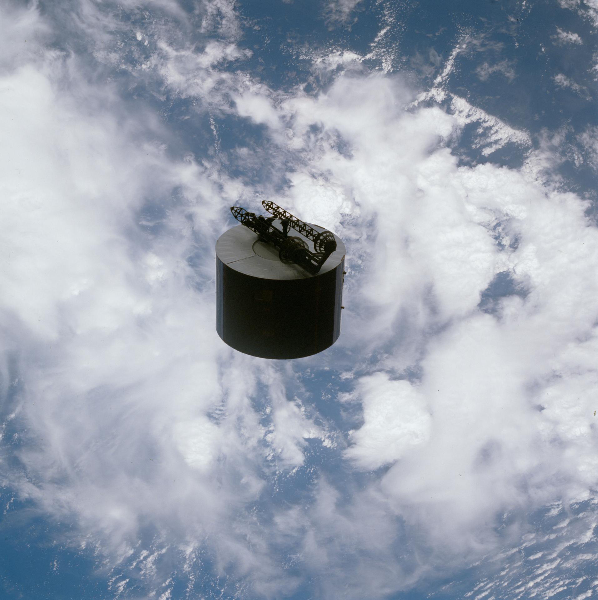 The cylindrical Syncom IV satellite is seen after deployment with the clouds and oceans of Earth visible behind it.