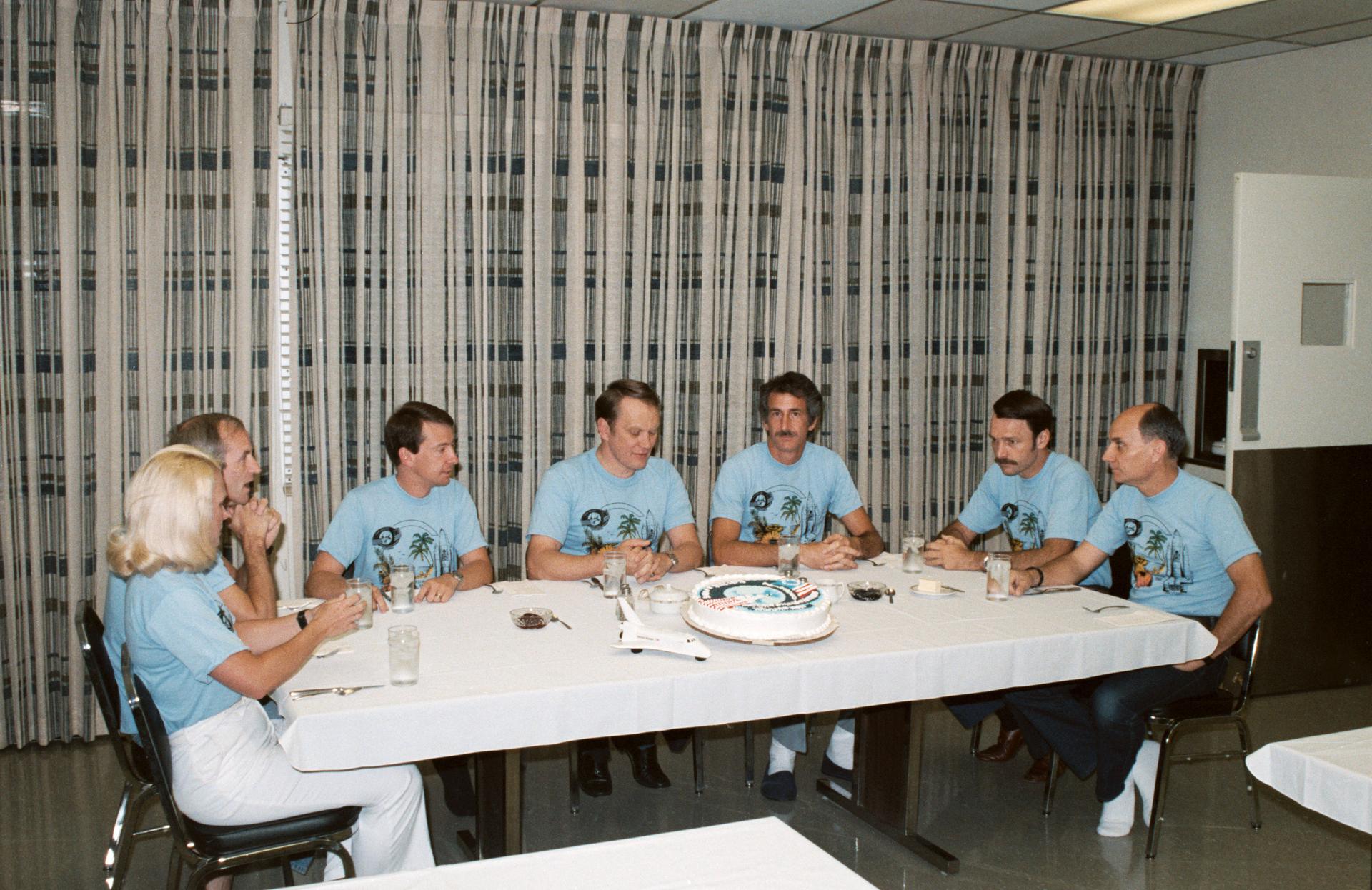 The 7 members of the STS-51D crew sit at a rectangular table for breakfast. A large cake is in the middle of the table next to a small model of a space shuttle.