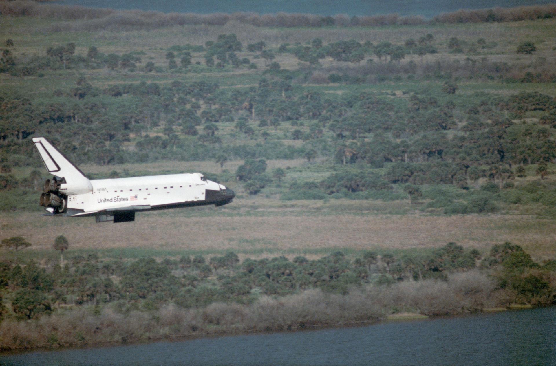 Space Shuttle Discovery prepares to land in Florida. Trees and water can be seen behind it.