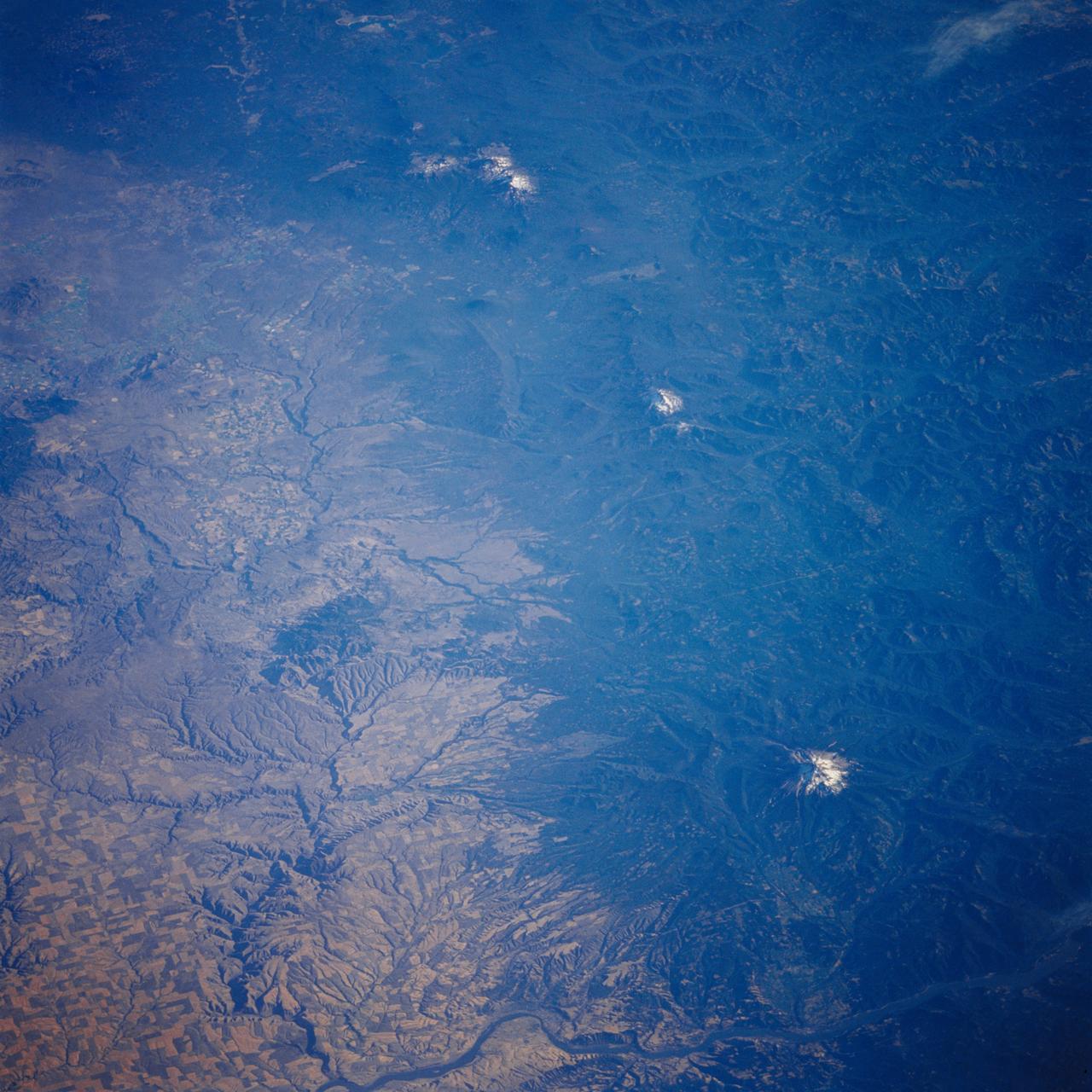 51F-37-097 (29 July-6 Aug 1985) --- The snow capped peaks of the Oregon Cascades are clearly seen.  From bottom to top we see Mount Hood, Mount Jefferson, and the Three Sisters volcanos.  The Columbia River is at the bottom.  The Deschutes River system and canyon, the scene of railroad wars nearly a century ago, is at the left side.  The Cascades make a very distinct rain shadow between the moist forests to the right and the  semiario lands to the east (left) of these great mountains.