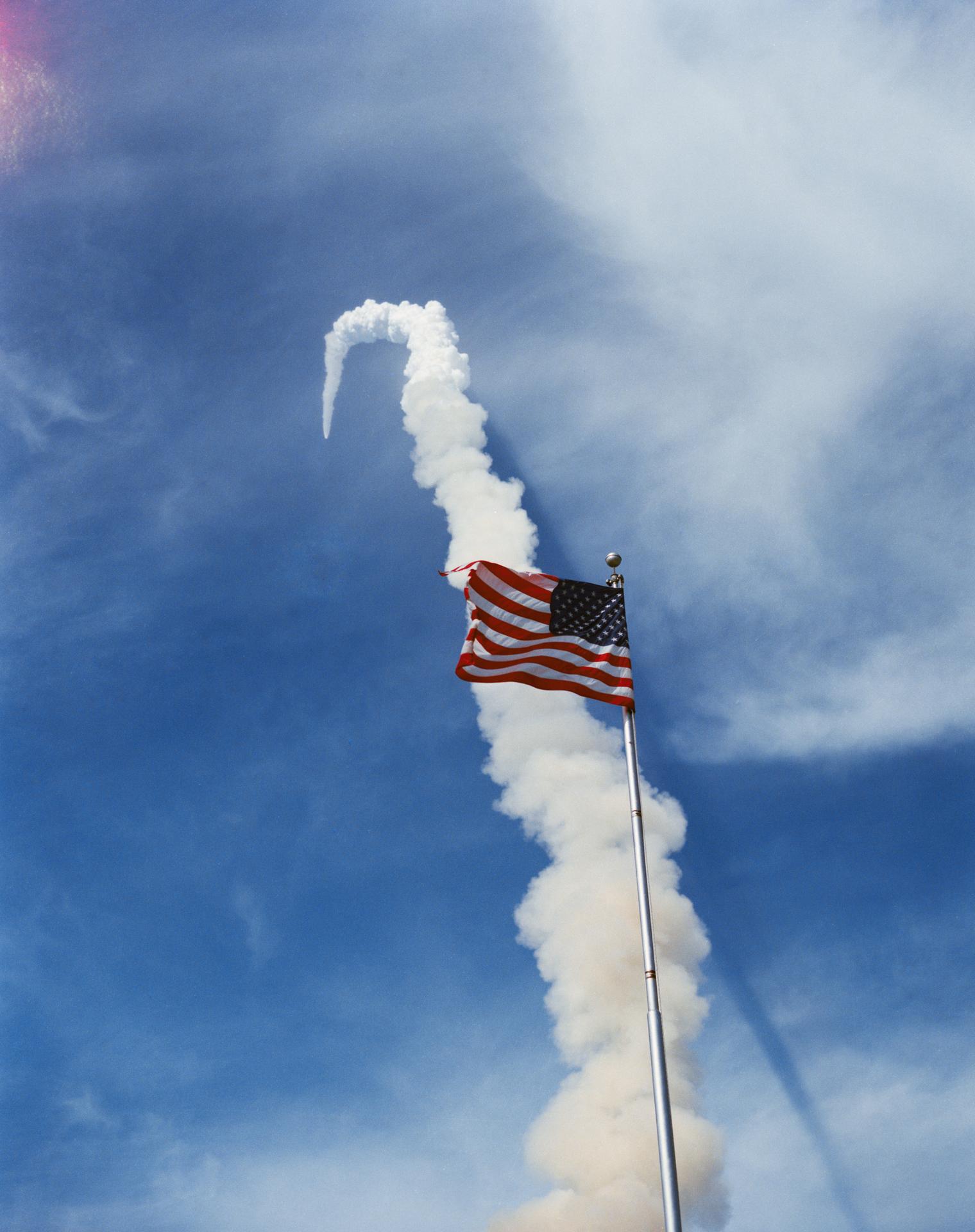 Trail of exhaust left after STS-51F launched into space. An American flag in the foreground overlays the trail