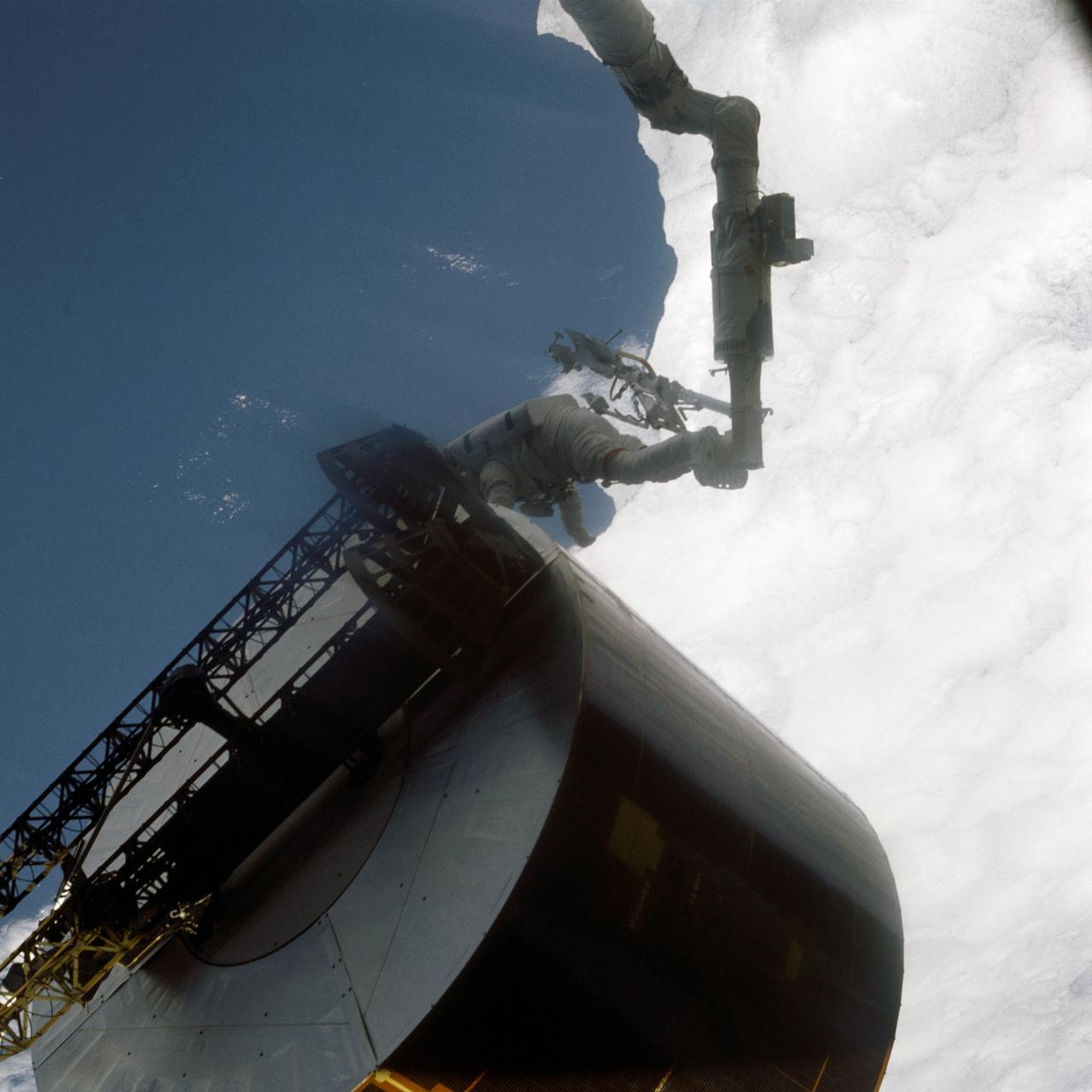 51I-44-014 (31 Aug-1 Sept. 1985) --- This photograph is one of a series of six covering extravehicular activity (EVA) which were released by NASA on Sept. 4, 1985. Here, astronaut James D. van Hoften, dwarfed by the large satellite, moves in for initial contact. Astronaut John M. (Mike) Lounge, out of frame inside cabin, maneuvers the remote manipulator system (RMS) arm to assist astronauts van Hoften and William F. Fisher. Photo credit: NASA